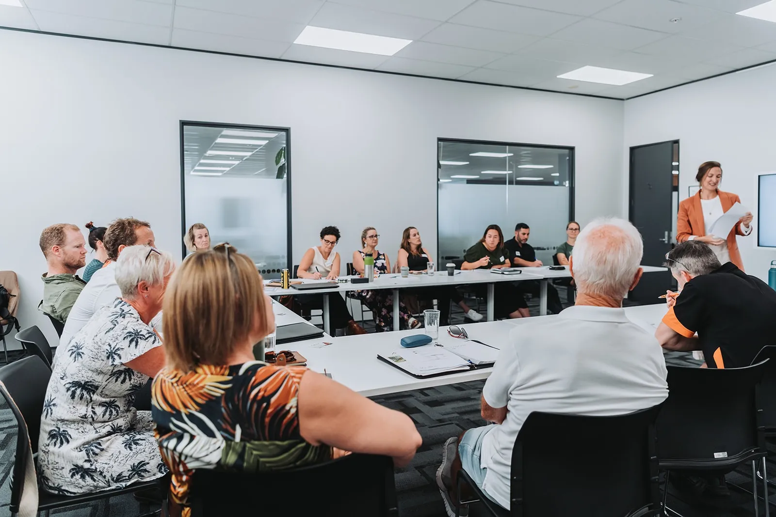 Group of adults sitting around large tables in a modern meeting room, listening to a woman standing and speaking while holding papers.