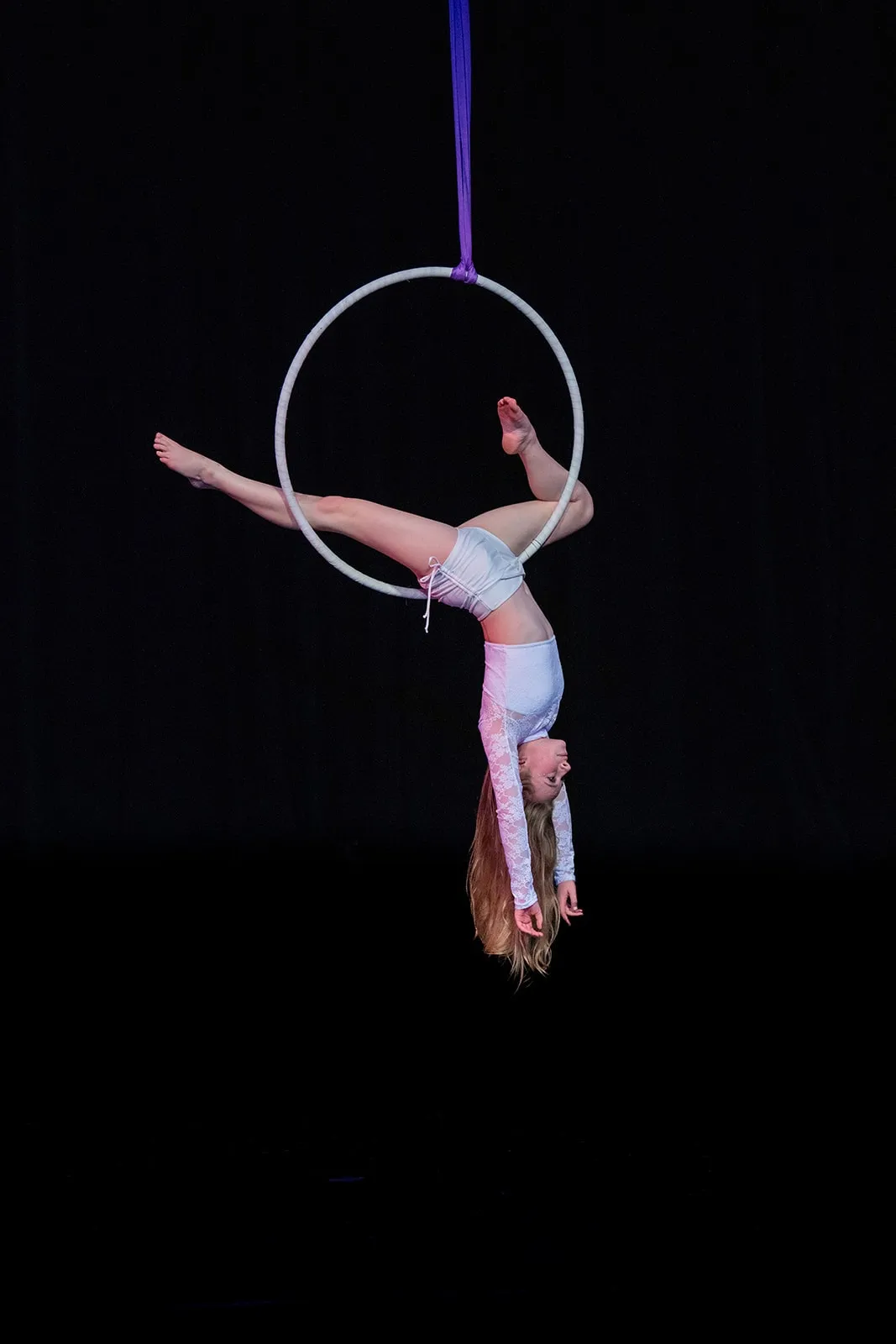 A female aerialist in white costume performing an upside-down pose on a suspended hoop against a black background.