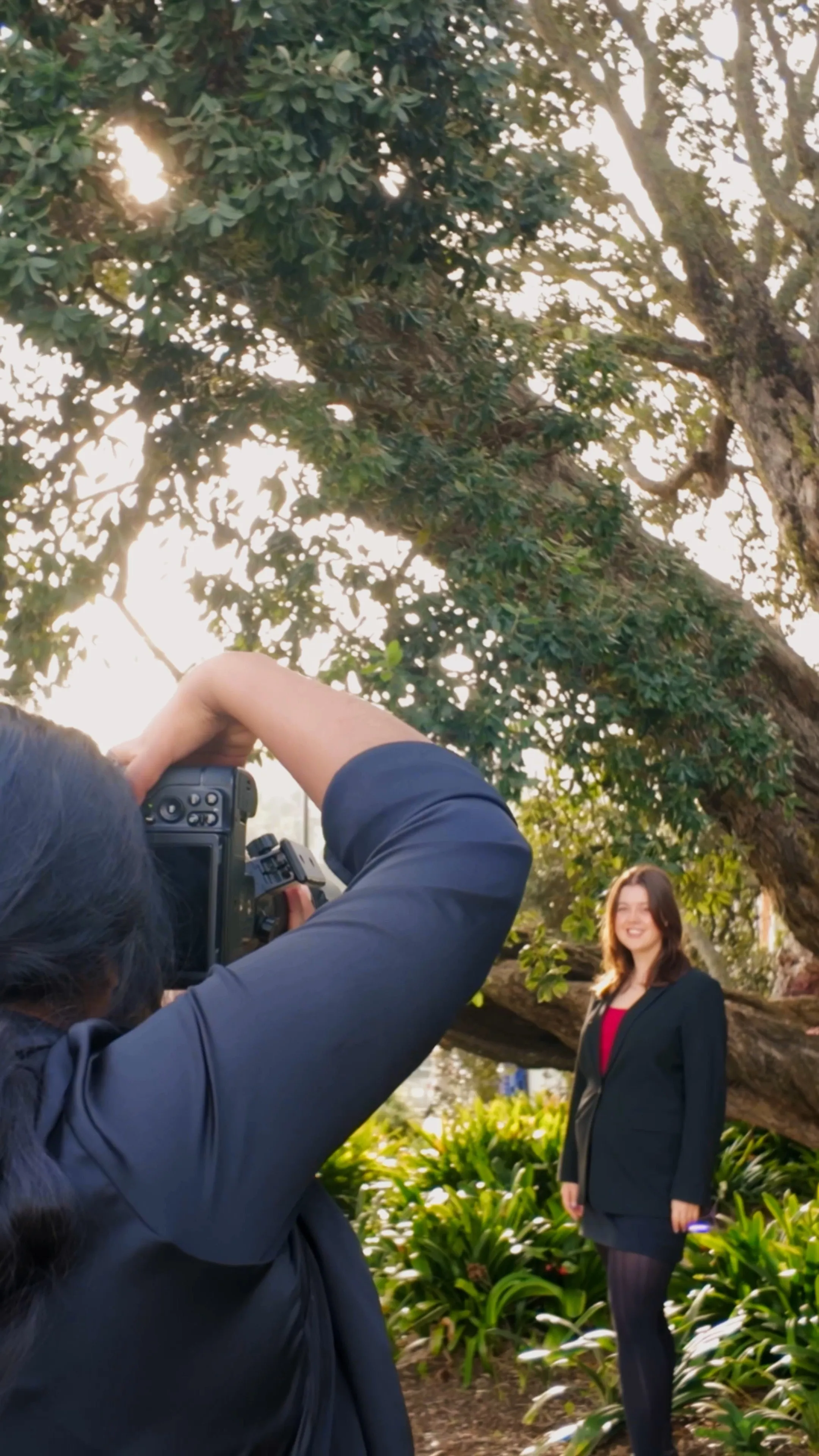 Photographer in a black jacket taking a photo of a smiling woman standing in a garden with green plants and a large tree. Corporate headshot photo session | Photography by Nimmy | Whangarei Photographer