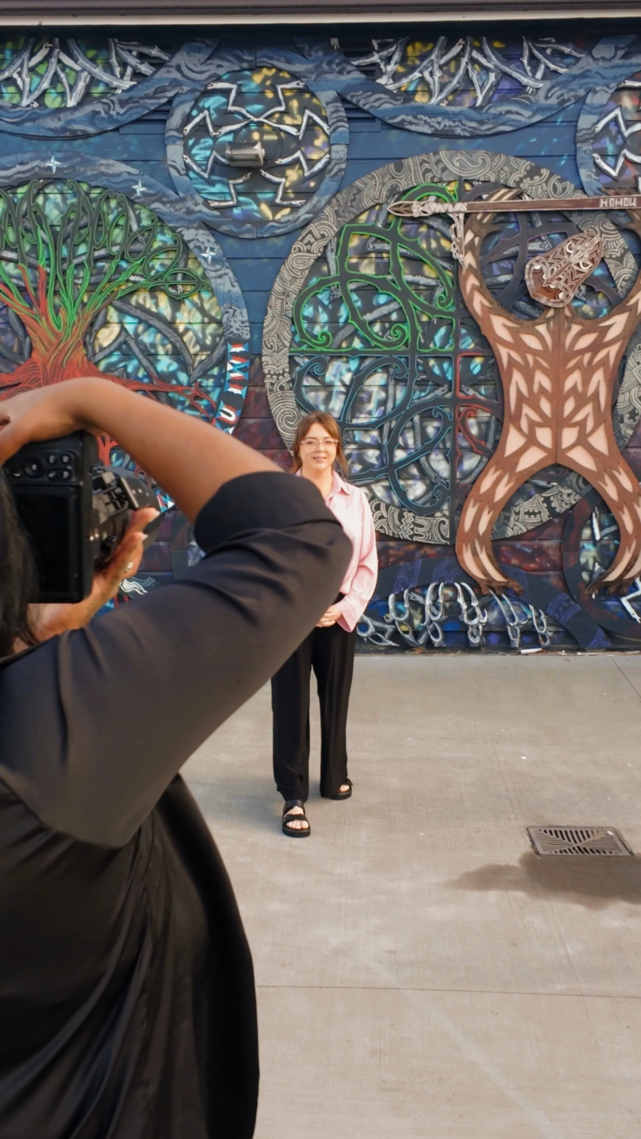 Photographer taking a photo of a woman standing in front of a colorful mural featuring a tree and an abstract figure holding a spear. Commercial headshot photo session Whangarei | Photography by Nimmy | Whangarei Photographer