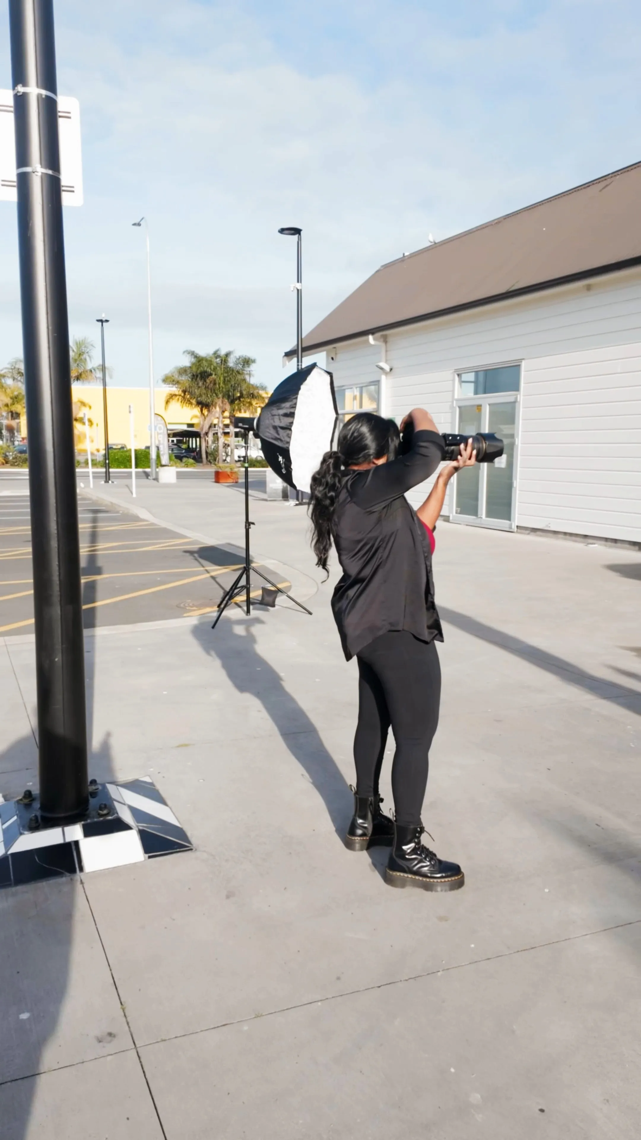 Photographer with long black hair wearing black clothes and boots, taking photos outdoors near a white building with a lighting setup beside her.