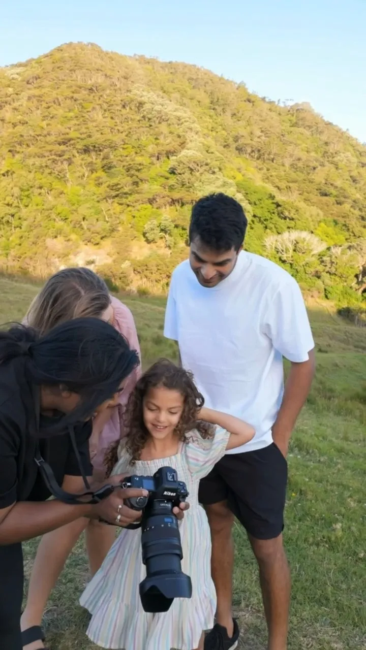 A group of four people outdoors looking at photos on a Nikon camera, standing on grass with a tree-covered hill in the background.