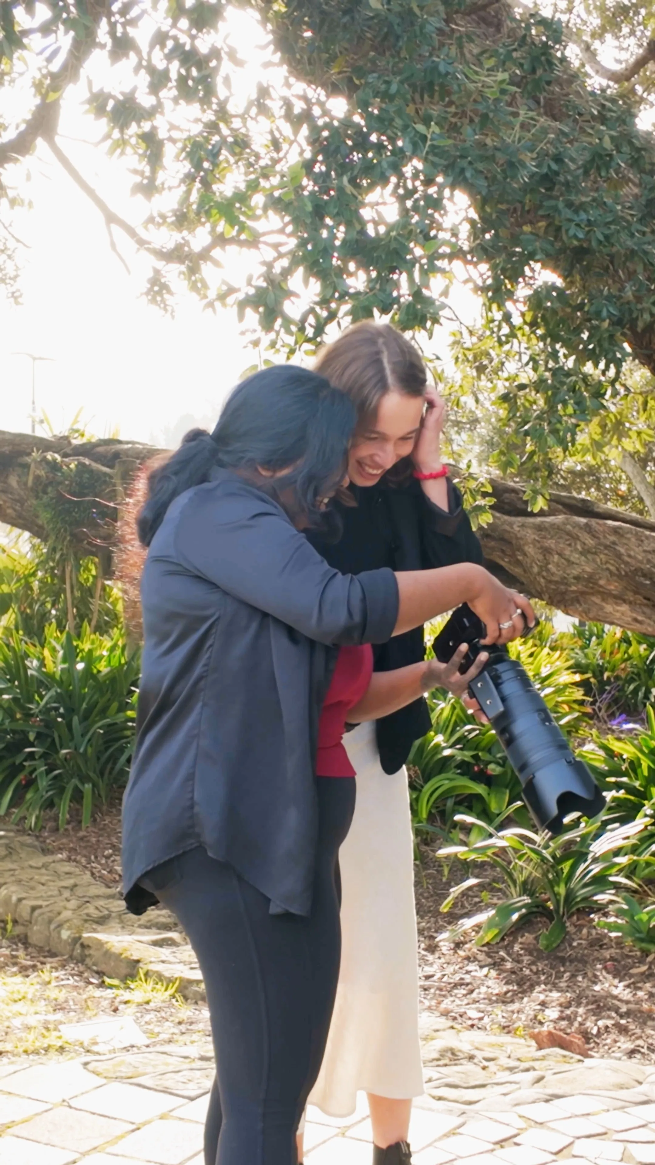 Two women outdoors reviewing photos on a digital camera under a large tree. Behind the scenes of a corporate headshot photography session | Photography by Nimmy | Whangarei Photographer