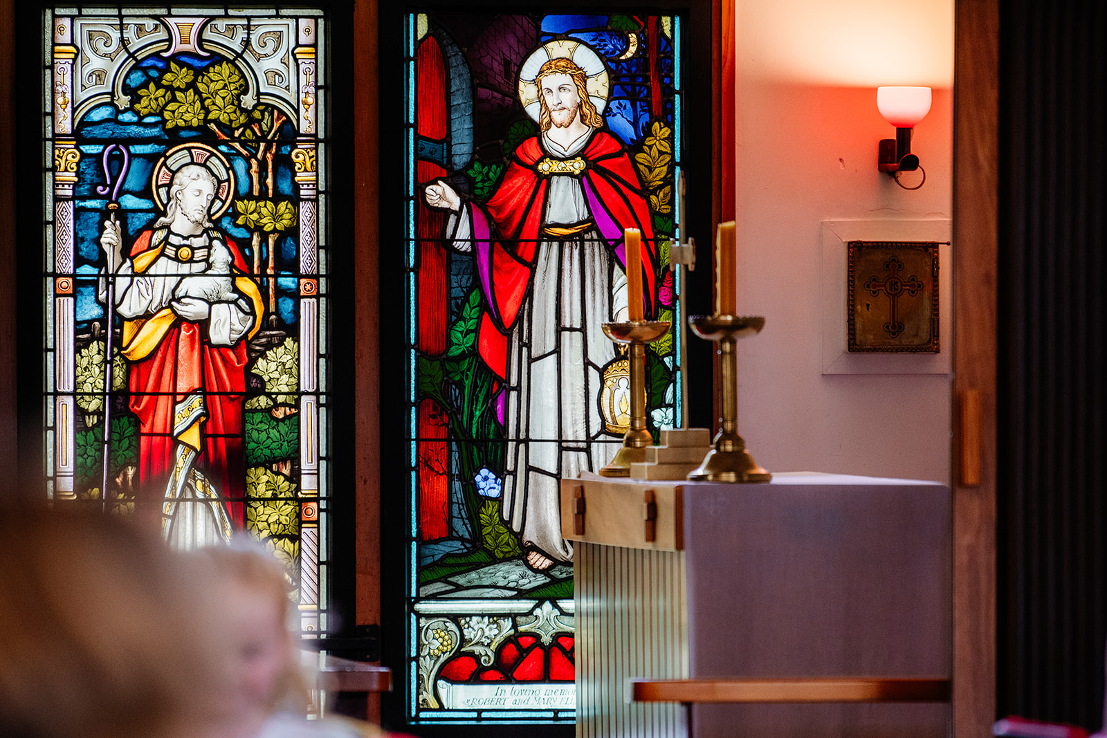 Two stained glass windows depicting Jesus in robes, alongside an altar with two lit candles and a wall lamp in a church interior; Photography by Nimmy | Whangarei Photographer