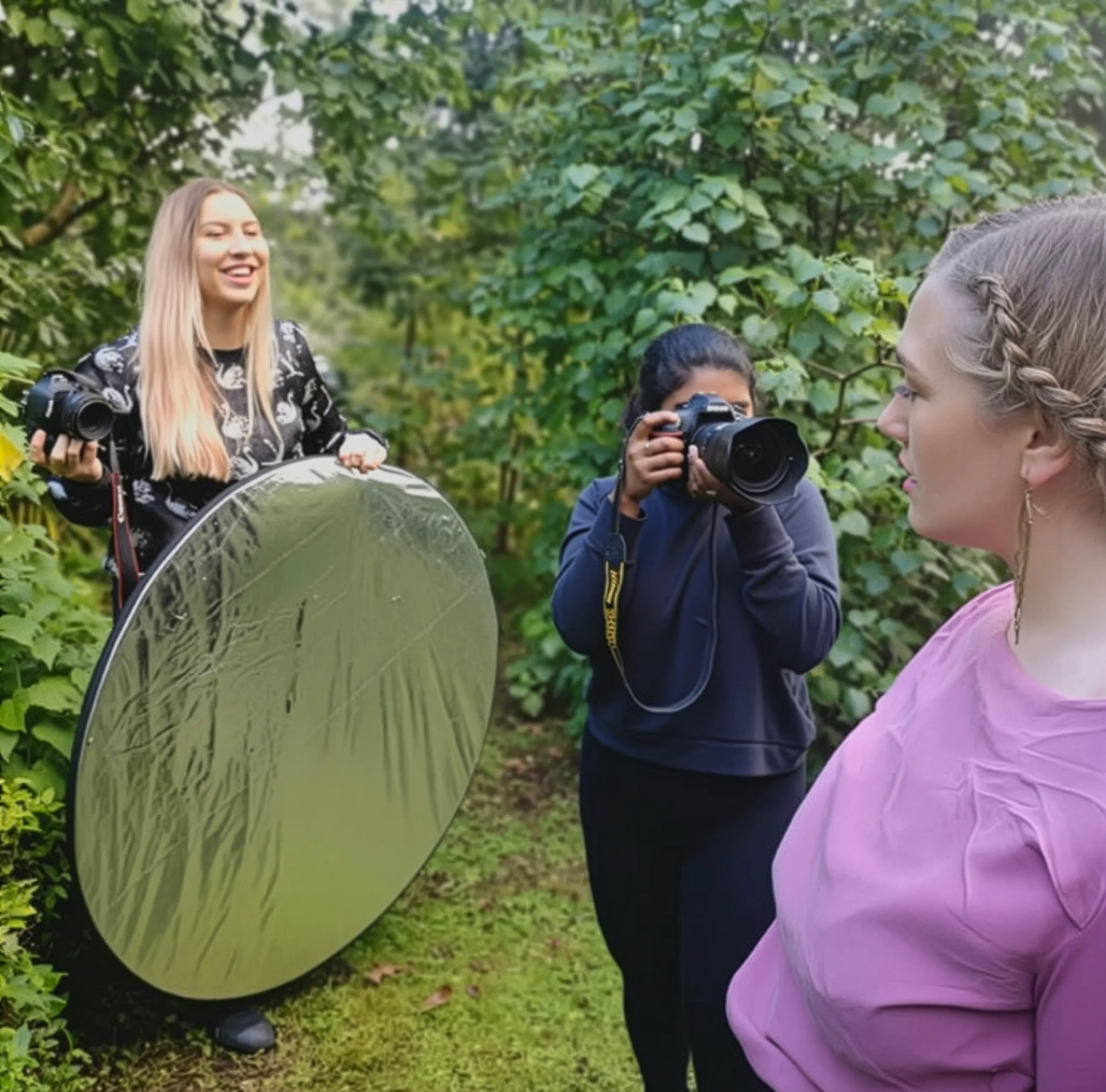 Photographer taking a picture of a woman in a pink top while an assistant holds a circular reflector outside among green foliage.