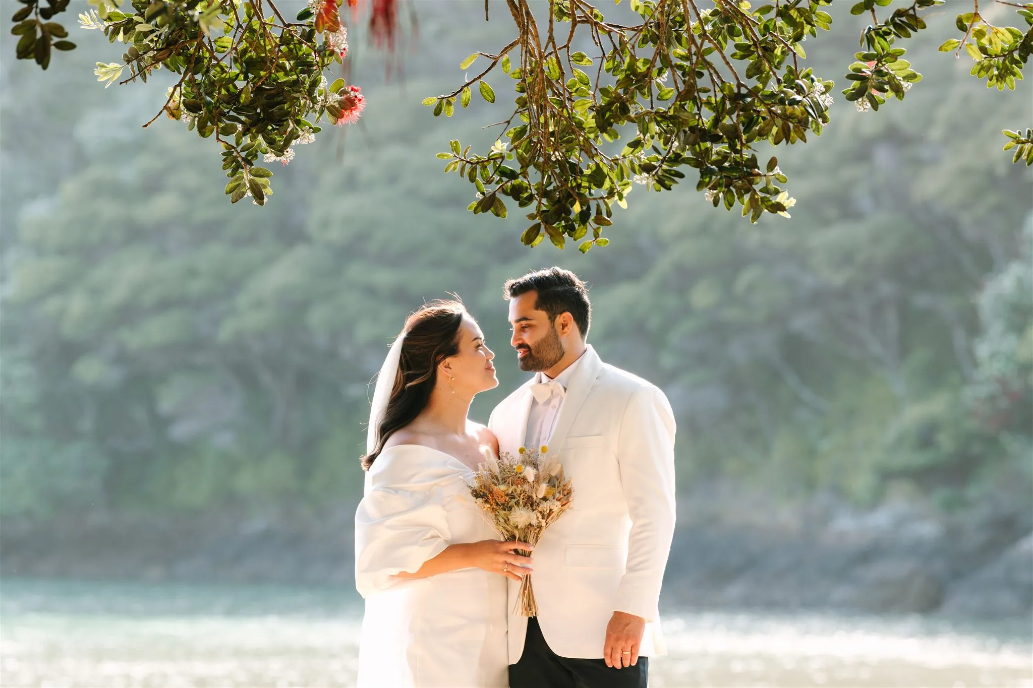 Bride and groom in white attire holding a bouquet, standing outdoors under leafy branches.