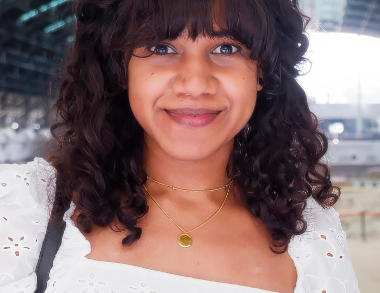 Smiling woman with curly hair, wearing a white eyelet top and a gold layered necklace.
