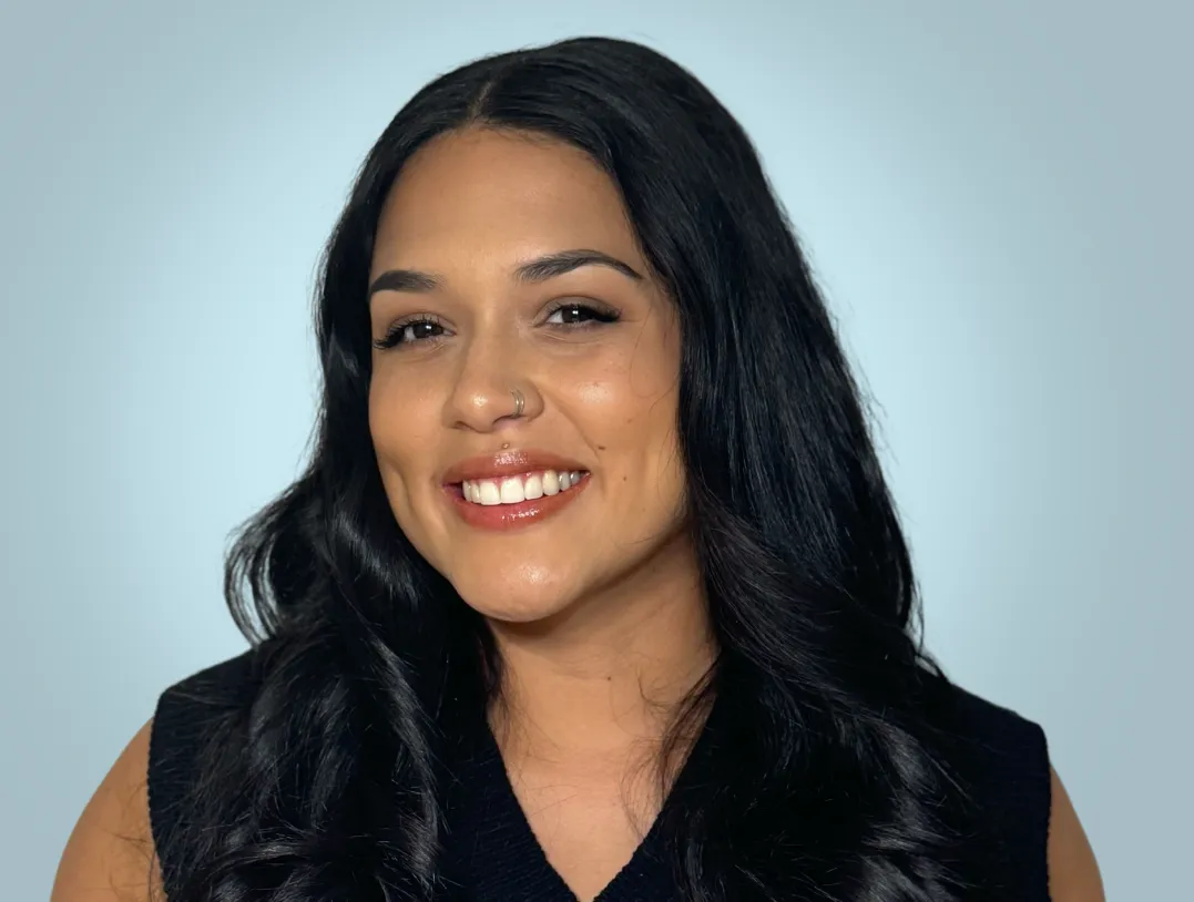 Smiling woman with long black hair, dimples, nose ring, and wearing a dark sleeveless top against a light blue background.