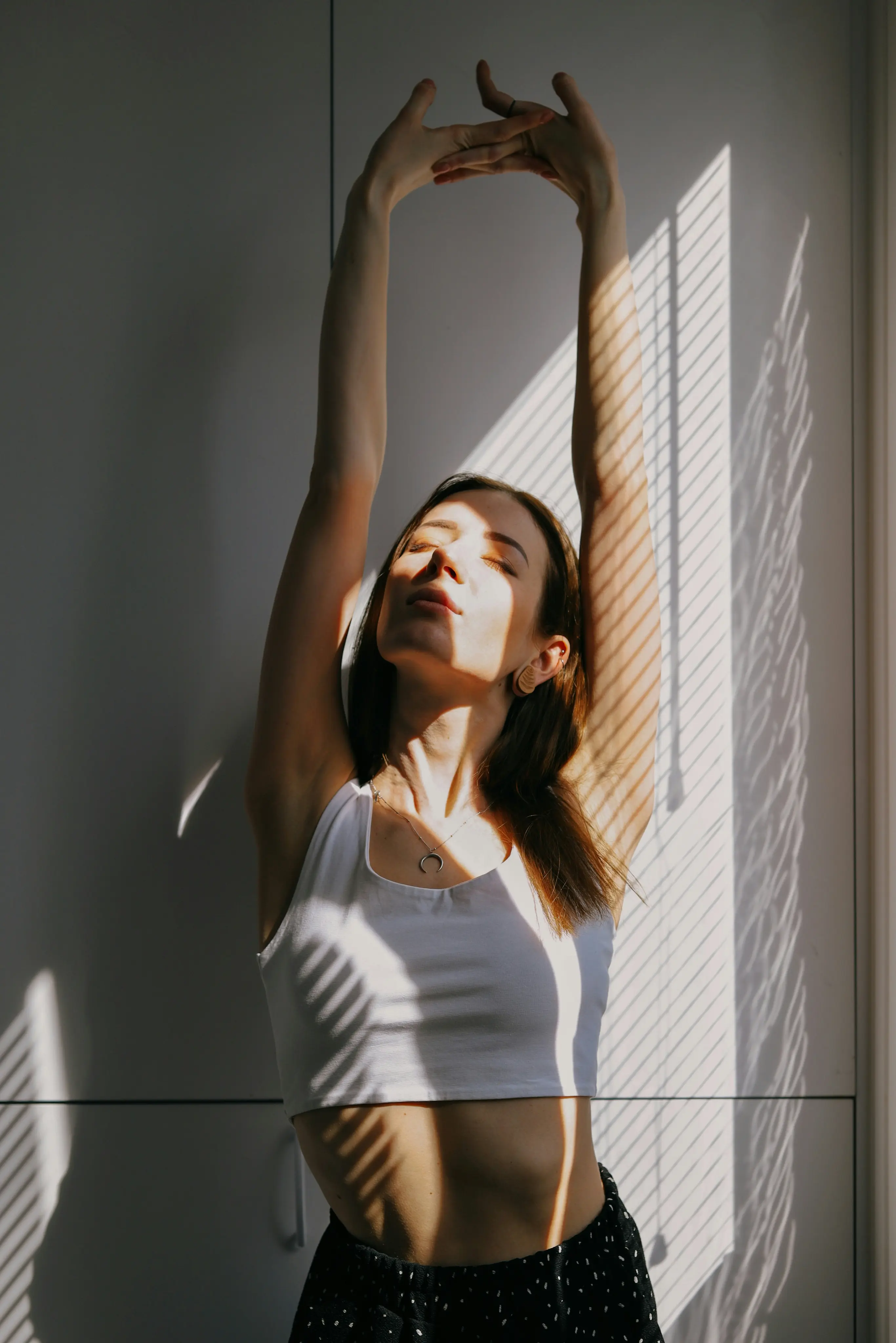 Woman stretching with eyes closed, sunlight casting striped shadows on her and the white wall behind.