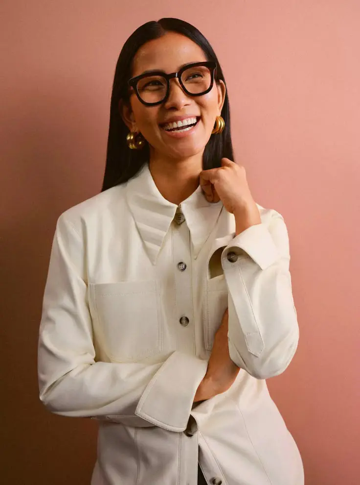 Smiling woman with long black hair wearing large glasses, gold hoop earrings, and a cream button-up shirt against a muted pink background.