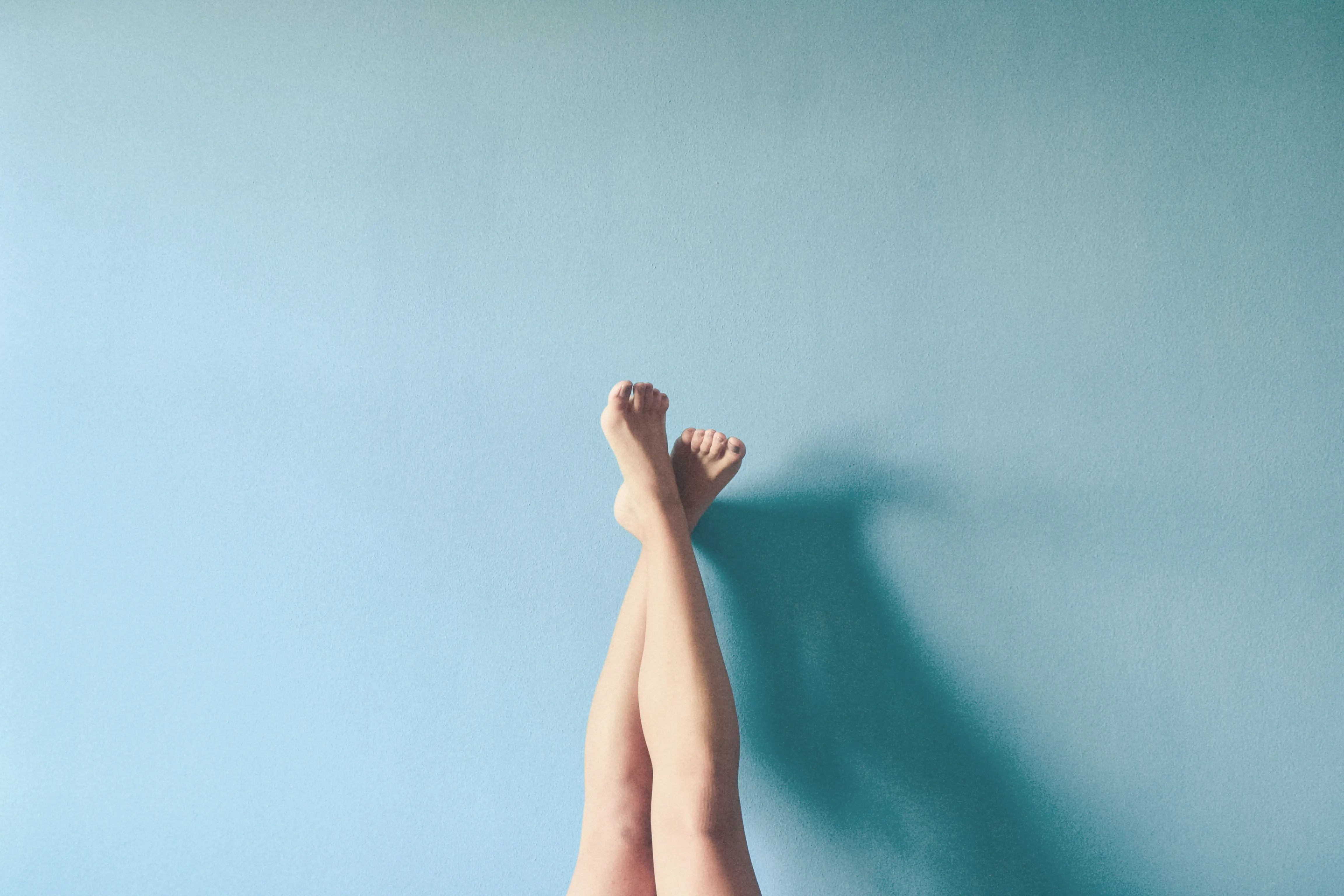 Pair of bare legs extended upward against a pale blue wall casting a soft shadow.