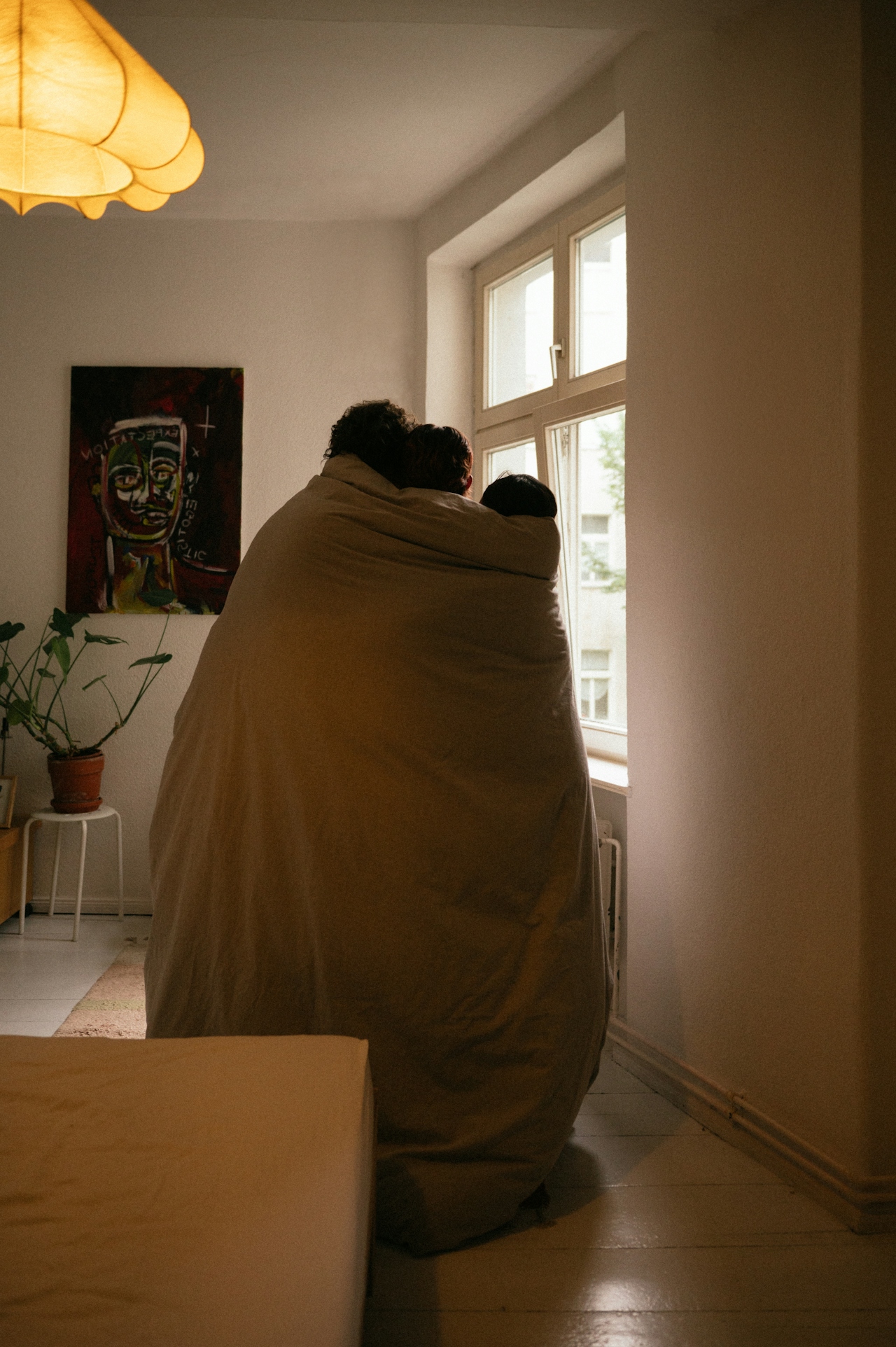 Three people wrapped together in a large blanket standing by a window in a cozy room.