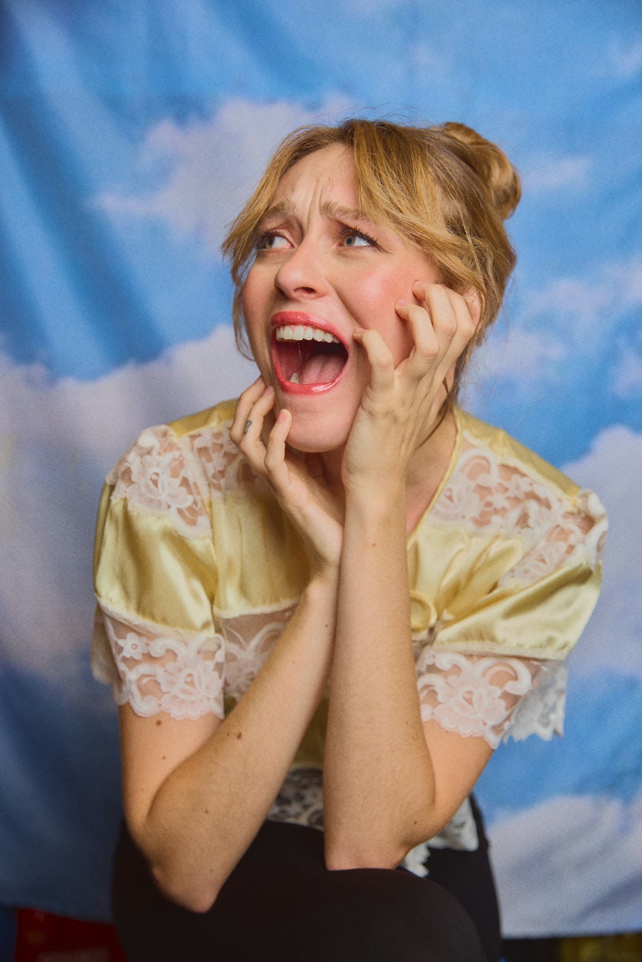 Blonde woman in a yellow lace dress with a distressed expression, holding her face and looking upward against a blue sky background.