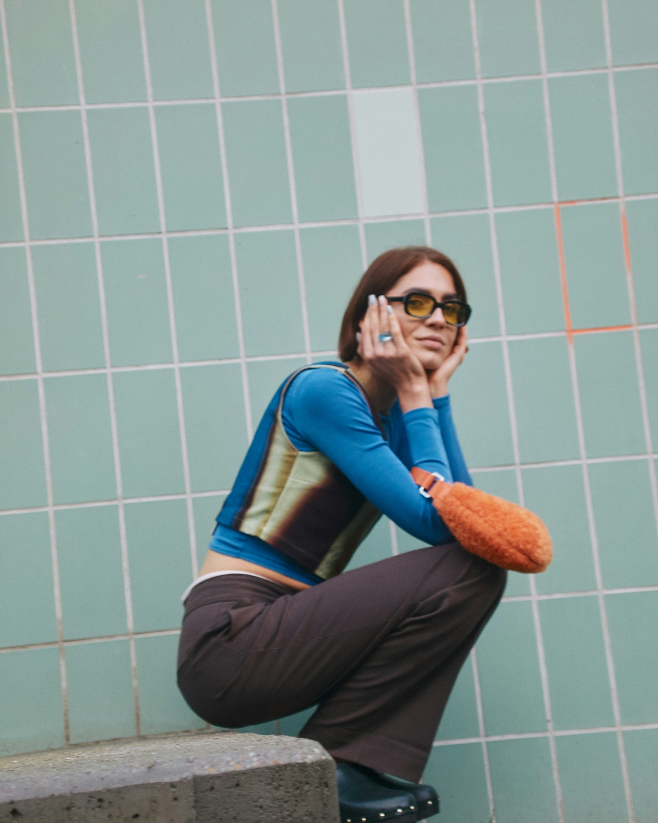 Woman in blue long-sleeve top, brown pants, and yellow tinted glasses crouching against a tiled green wall, holding her face with both hands.