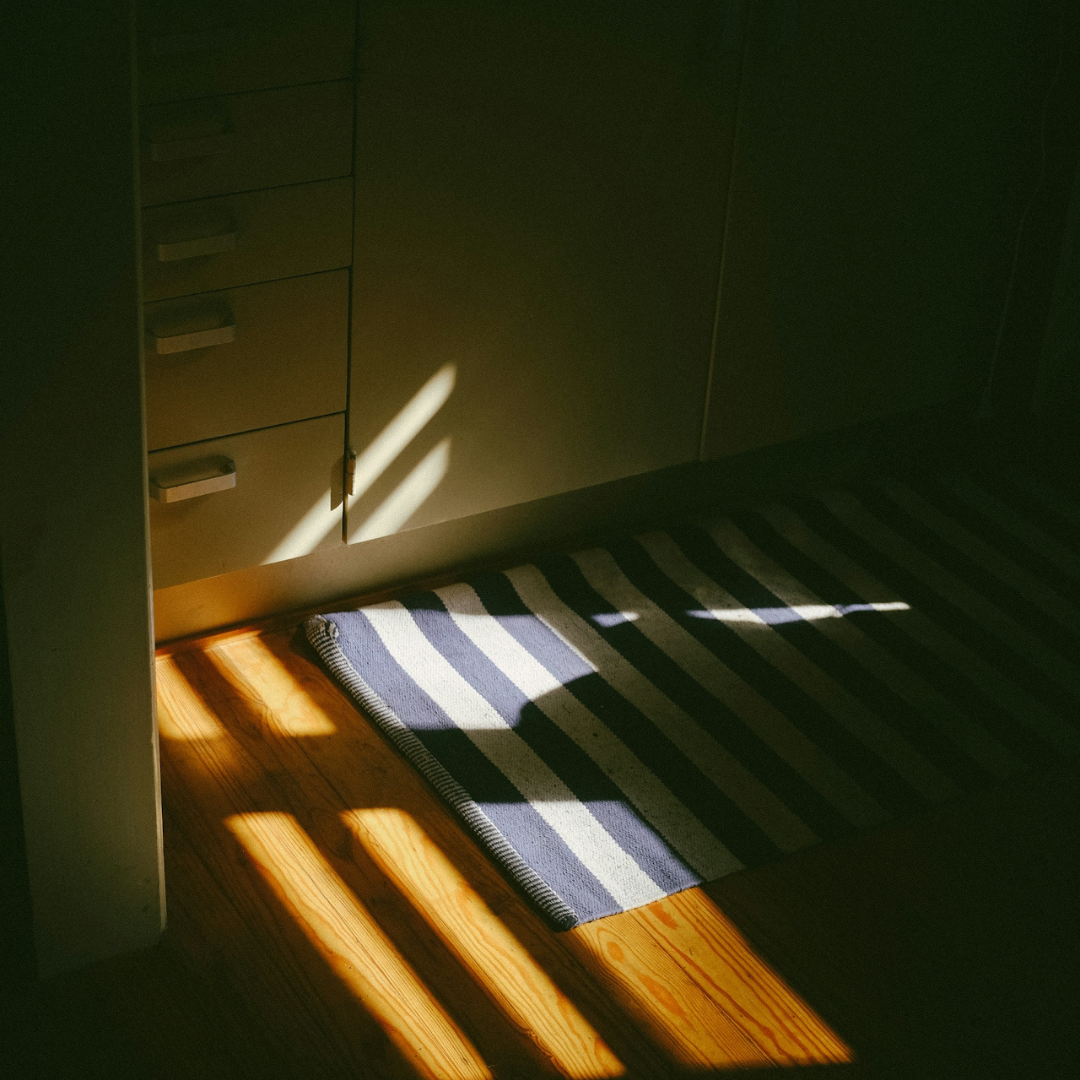 Sunlight casting shadows on a wooden floor and striped rug near a cabinet with drawers.