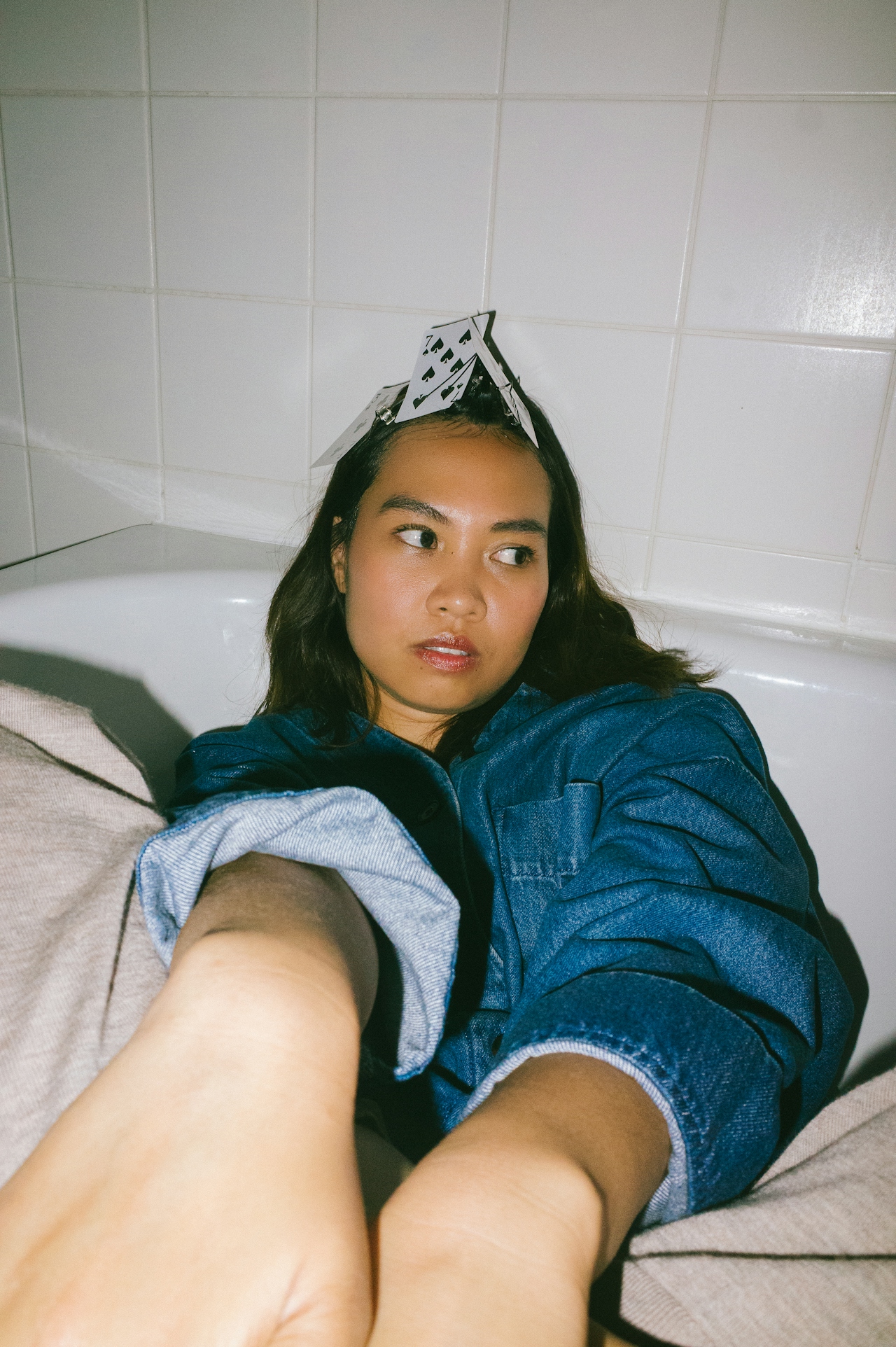 Young woman in a denim jacket sitting in a bathtub with playing cards arranged on her head.
