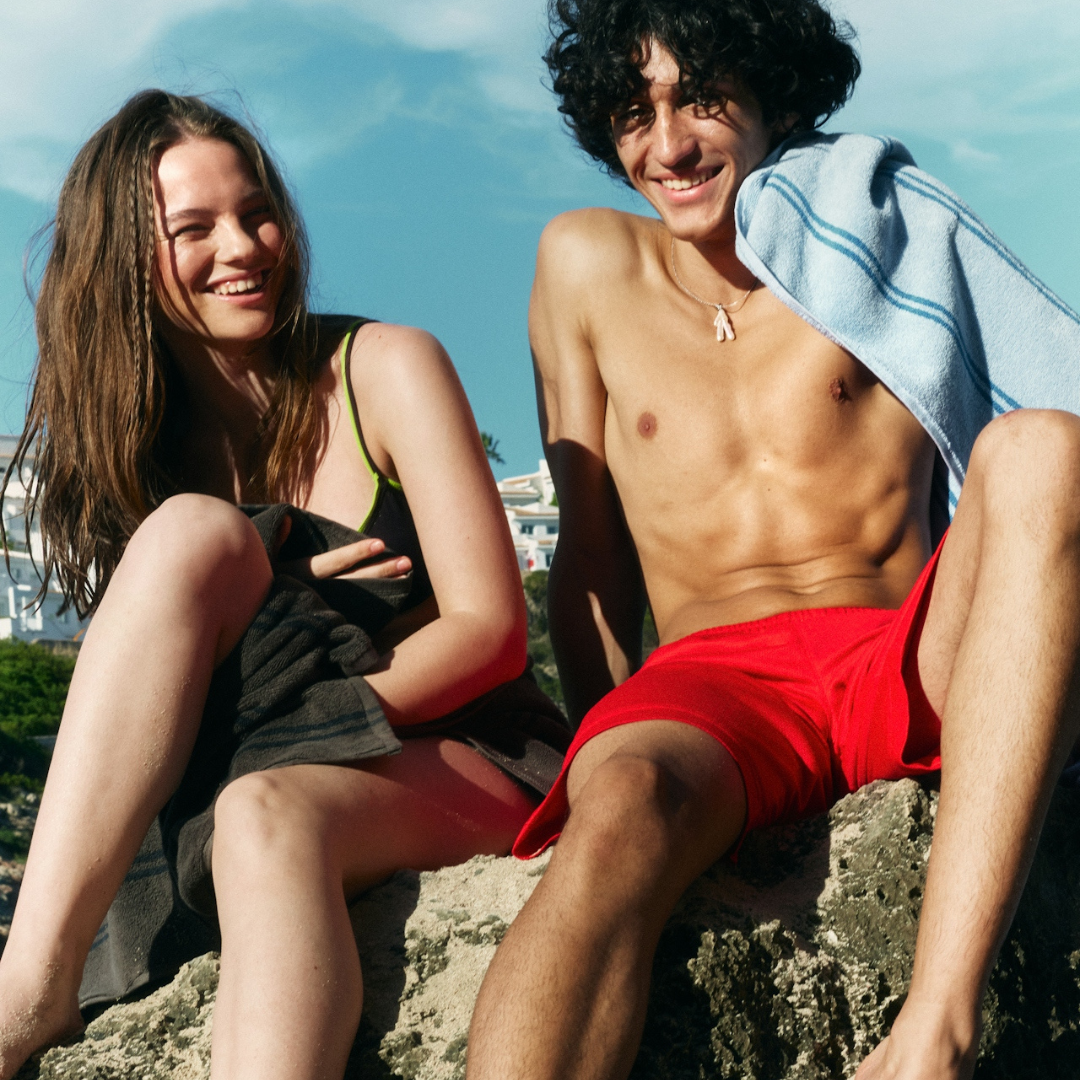 Smiling young man and woman sitting on rocks outdoors, wearing swimwear with towels, under a blue sky.
