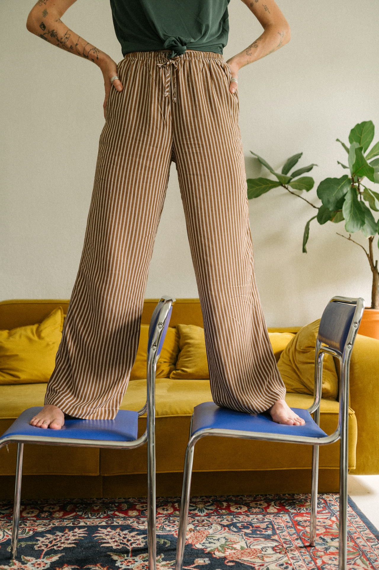 Person standing barefoot with legs apart on two blue chairs in a living room with a yellow couch and a large green plant.
