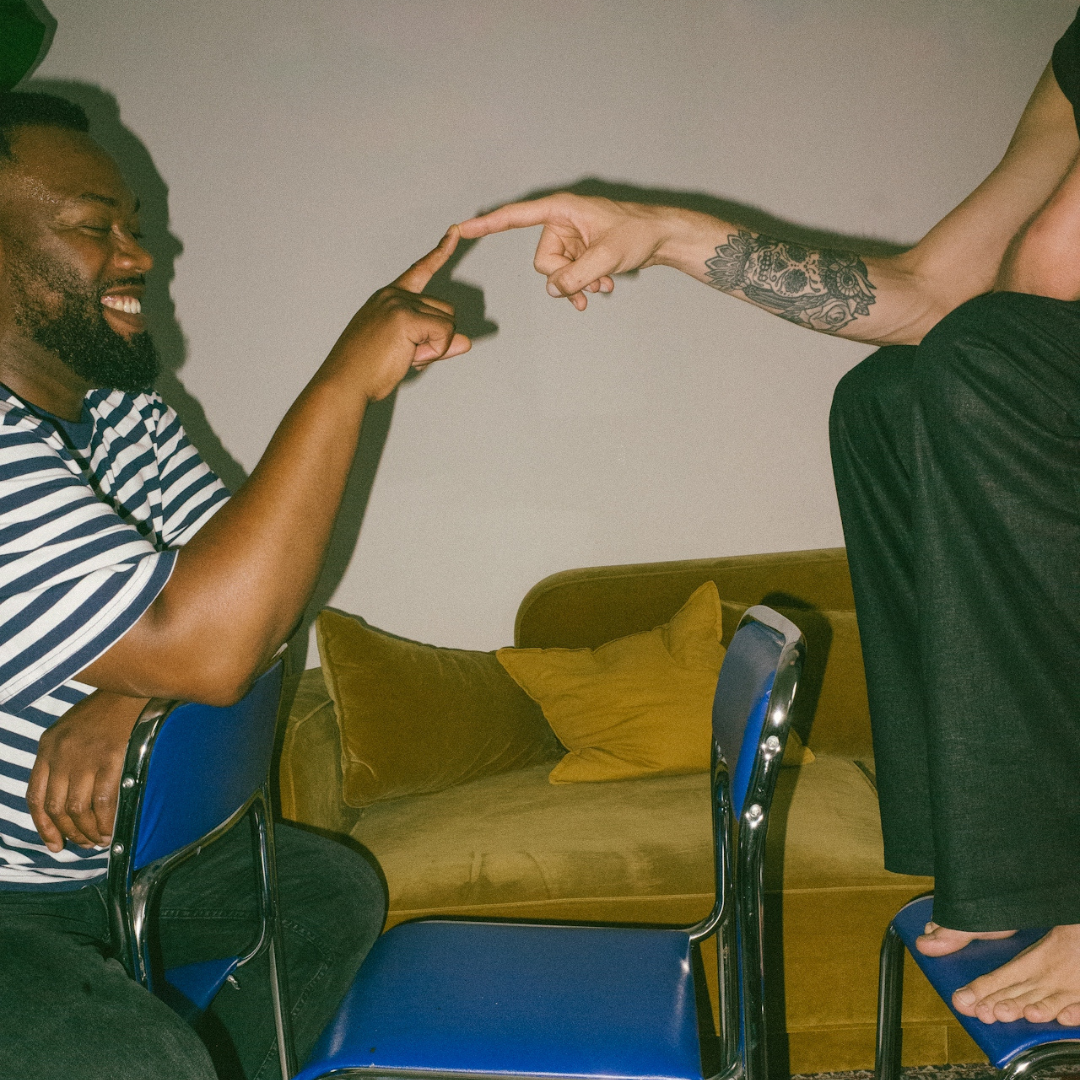 Two men smiling and touching their index fingers together while sitting on blue chairs near a yellow couch.