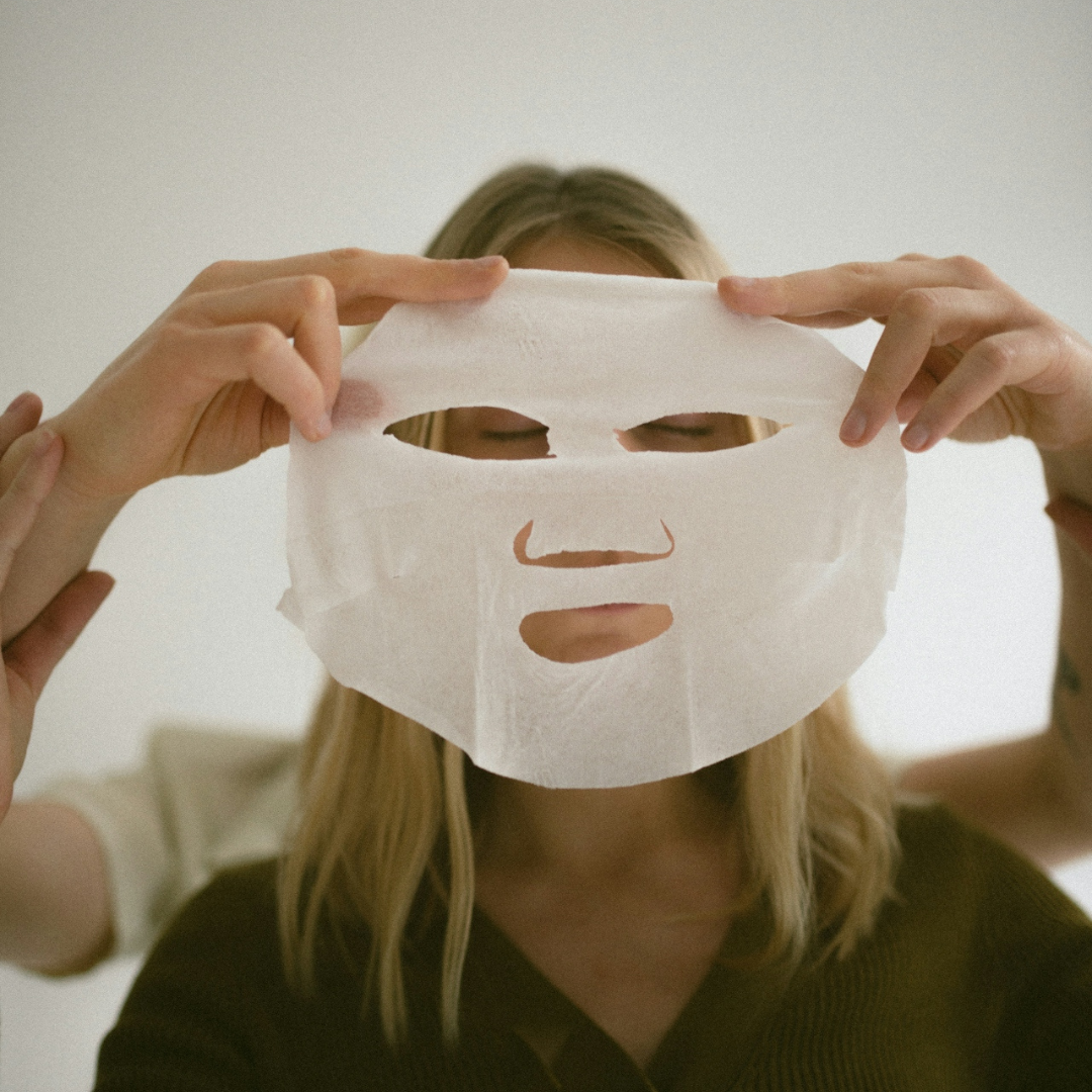 Person holding a white sheet face mask in front of a woman's face with closed eyes.