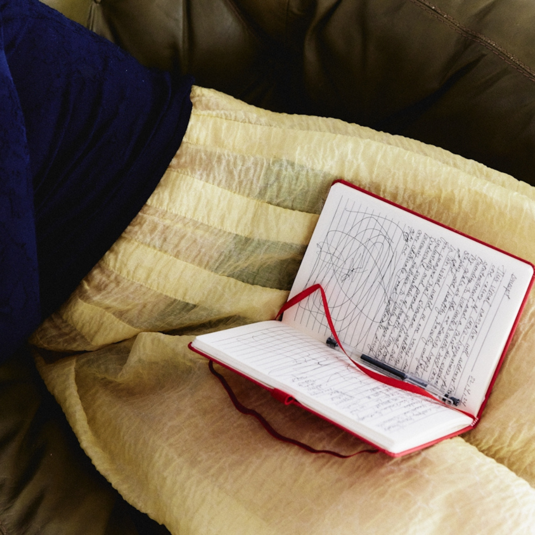 Open red notebook with handwritten notes and a black pen resting on a yellow striped blanket on a brown couch.