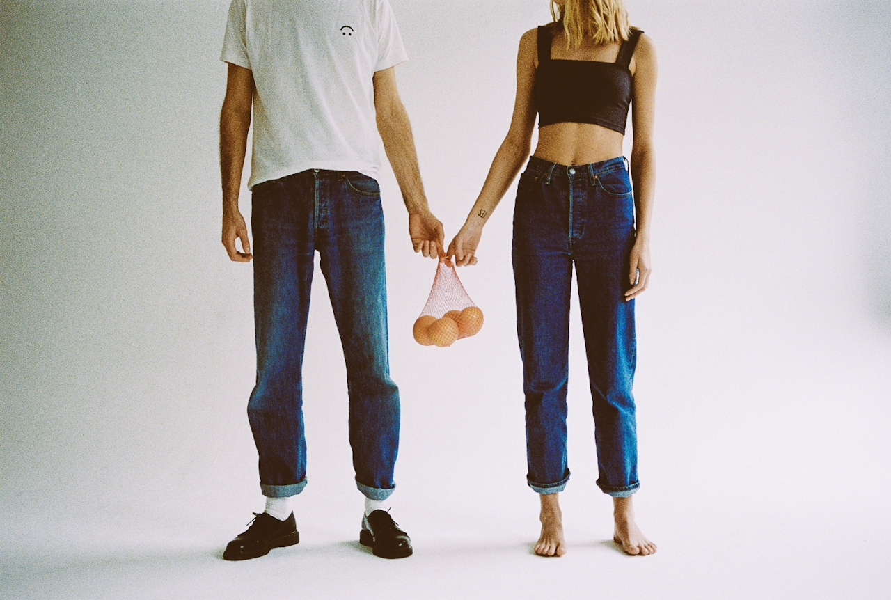 Man and woman standing side by side holding a small net bag of oranges together, facing forward against a plain background.