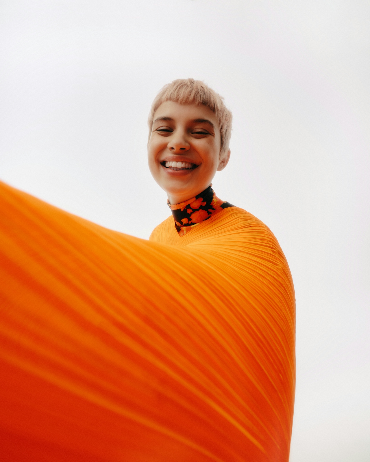 Smiling person with short light hair wearing a bright orange pleated garment against a white background.