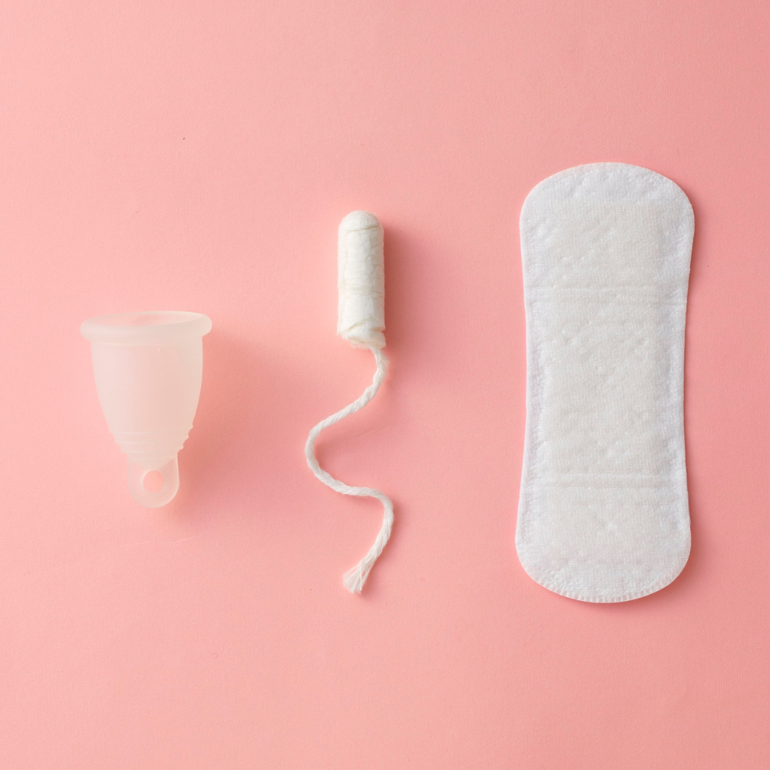 Three menstrual hygiene products on a pink background: a menstrual cup, a tampon with string, and a panty liner.
