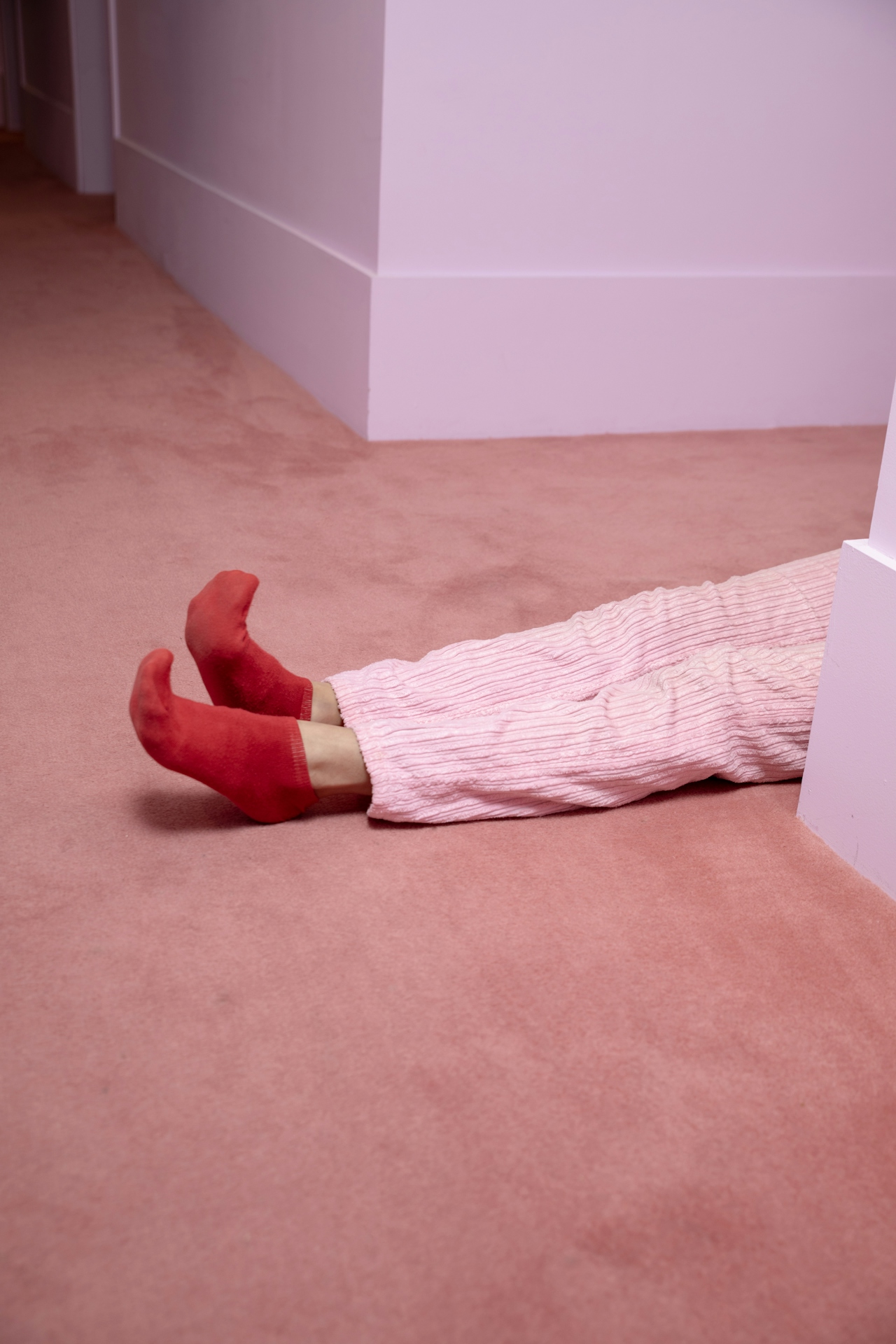 Person lying on a pink carpet floor wearing pink pants and red socks, partially hidden behind a wall corner.