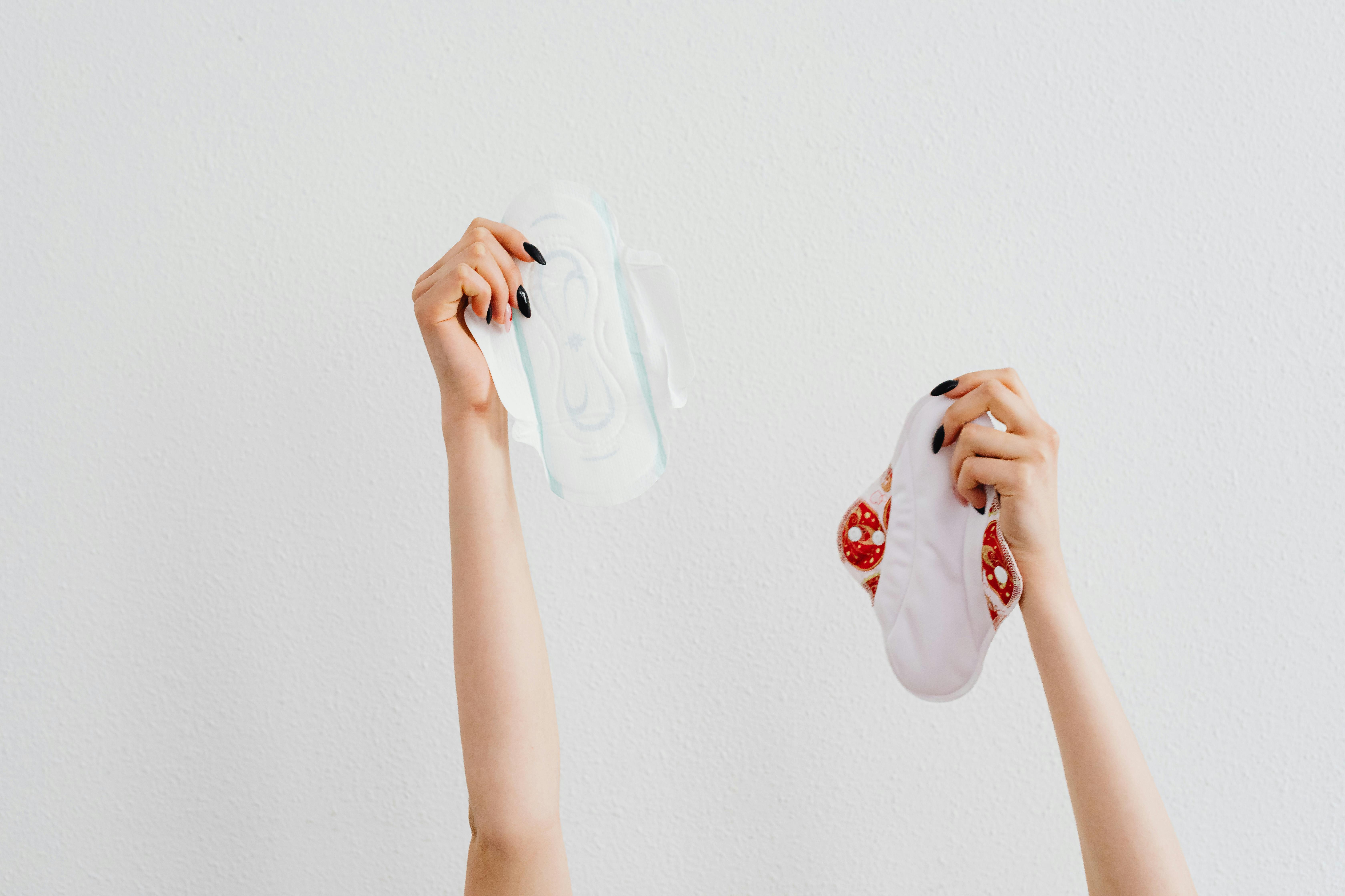 Two hands holding up two different sanitary pads against a white wall background.