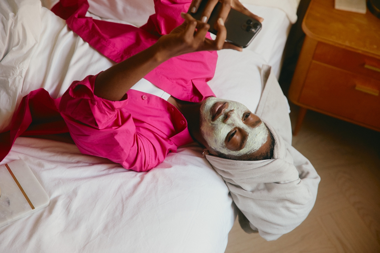 Person lying on a bed with a facial mask and a towel wrapped around their head, wearing a bright pink shirt and holding a smartphone.