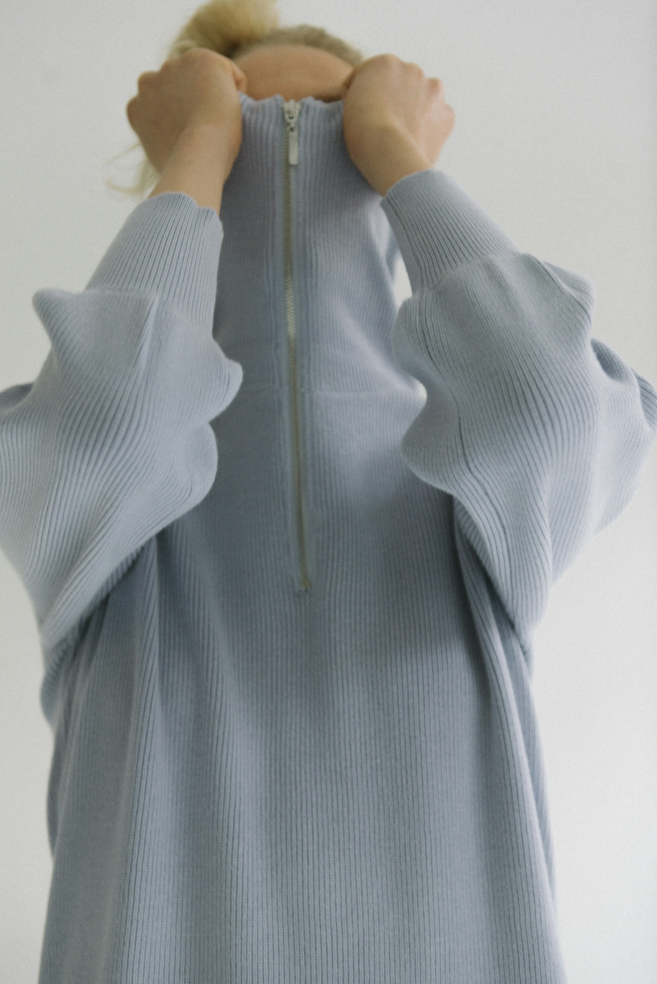 Person pulling the collar of a light blue ribbed sweater with a front zipper up over their face.