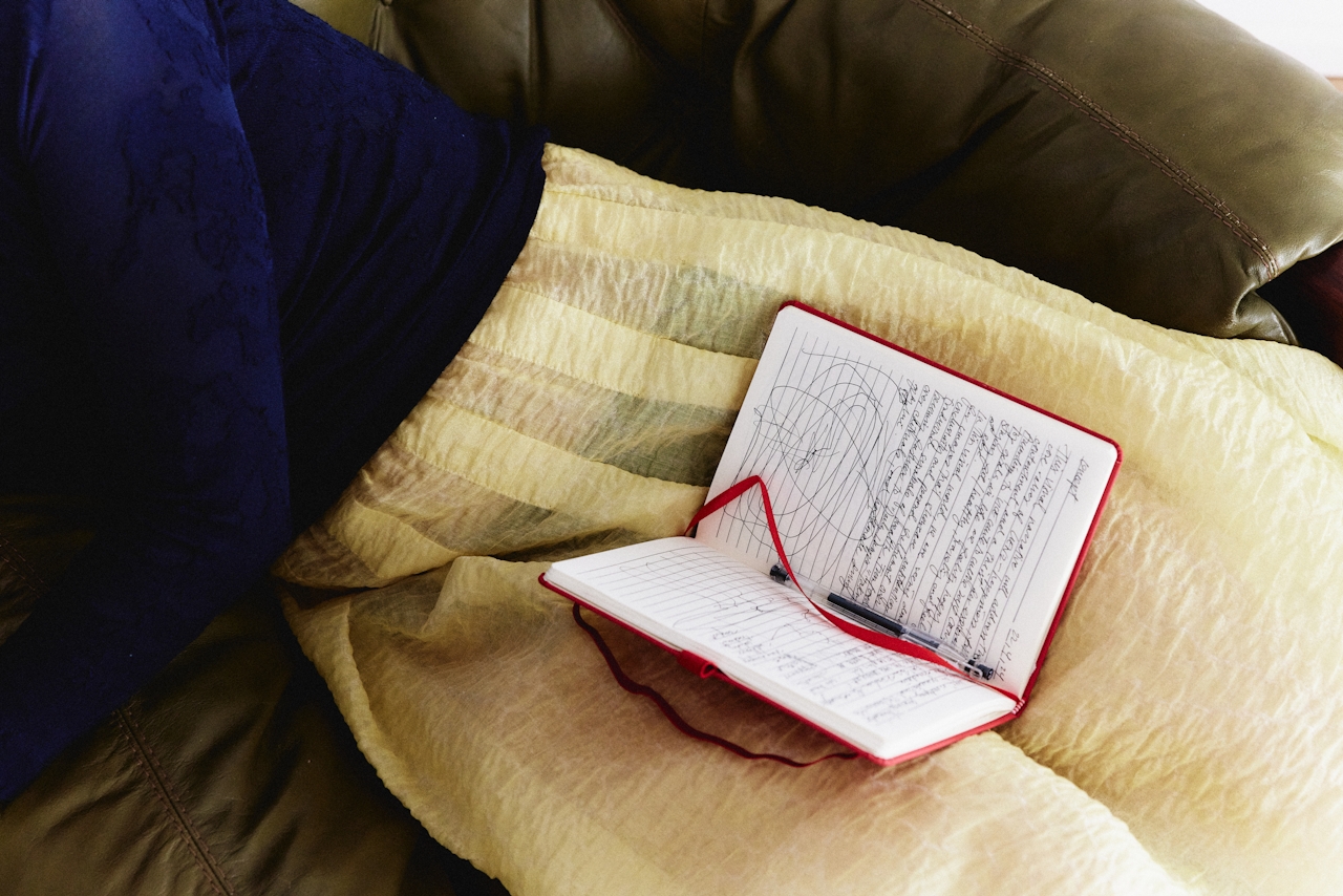Open notebook with handwritten notes and a pen resting on a yellow striped blanket over a brown leather couch.