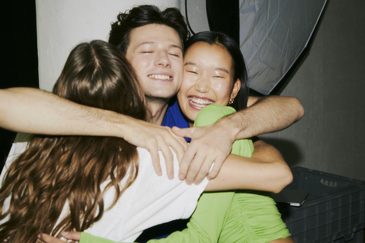 Three friends embracing tightly in a group hug, smiling with eyes closed, one wearing a bright green top.