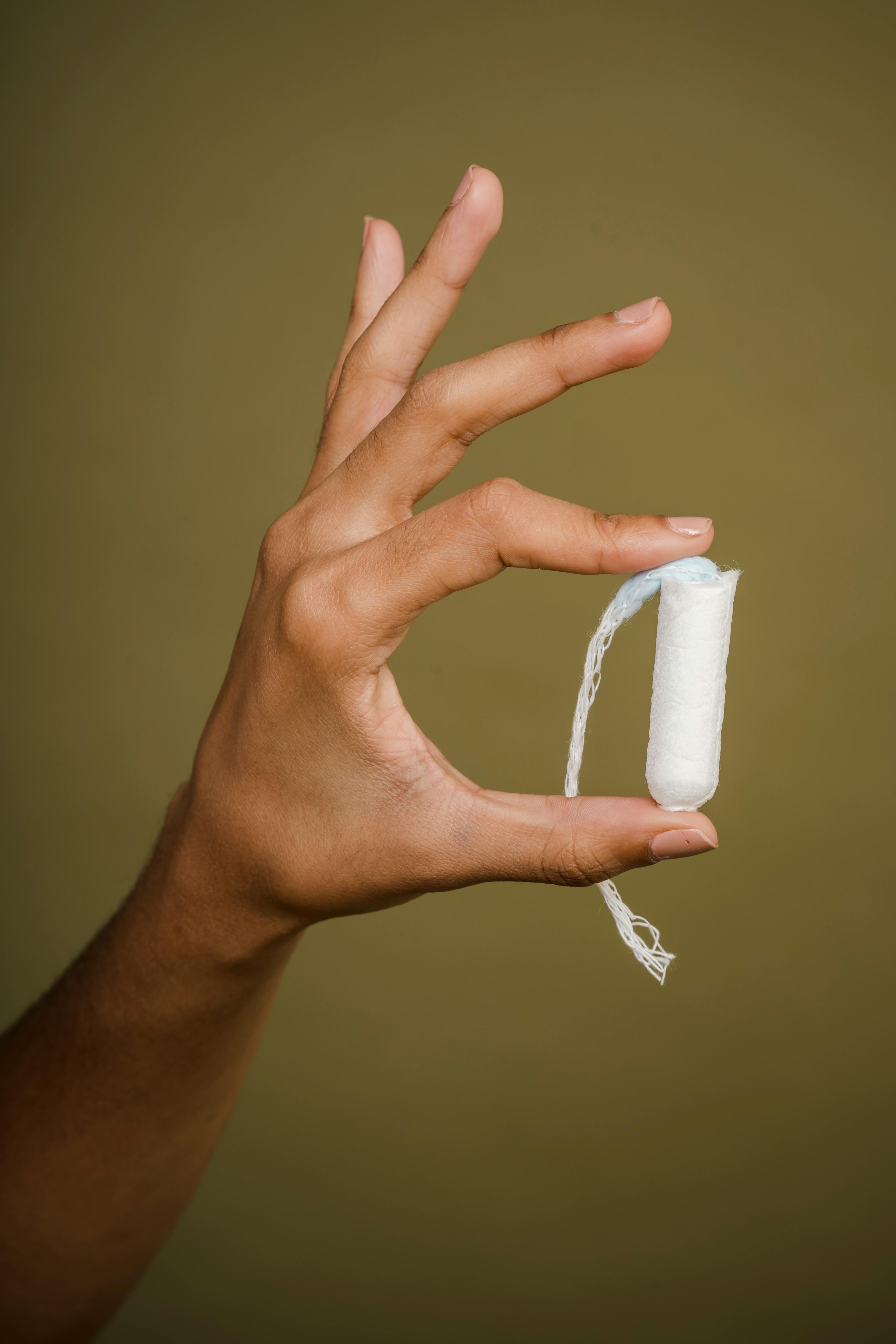 Hand with medium skin tone holding a white tampon with an extended string against a plain olive background.