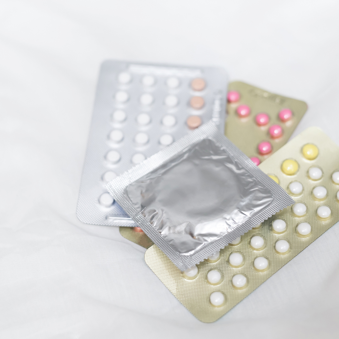 Blister packs of various contraceptive pills with one foil-wrapped condom on top, placed on a white fabric surface.