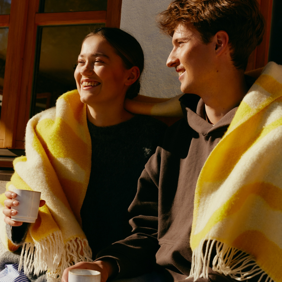 Smiling young man and woman sitting warmly wrapped in a yellow and white blanket, holding white mugs.