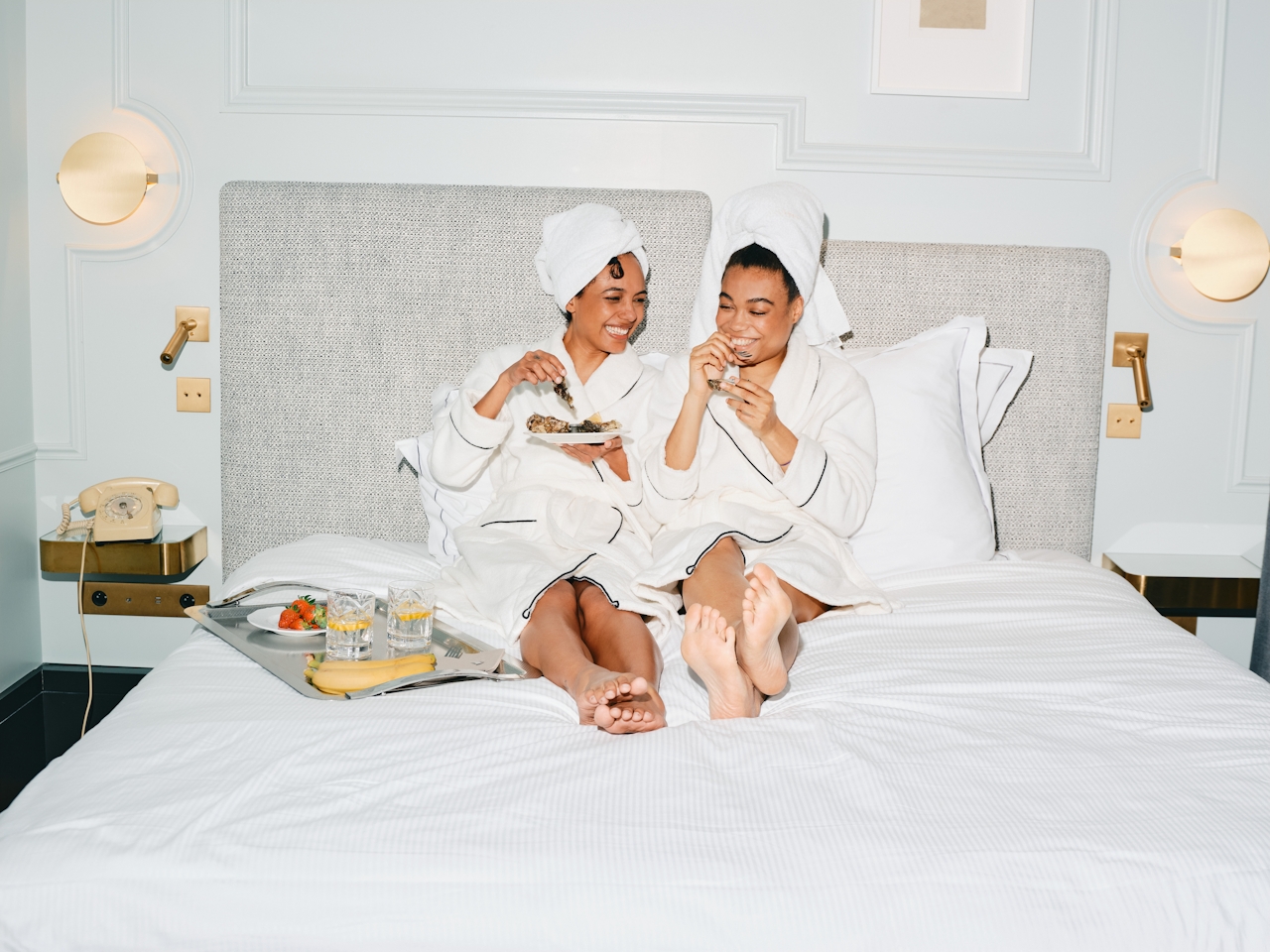 Two women in white bathrobes and towels on their heads sitting on a bed sharing breakfast and laughing.