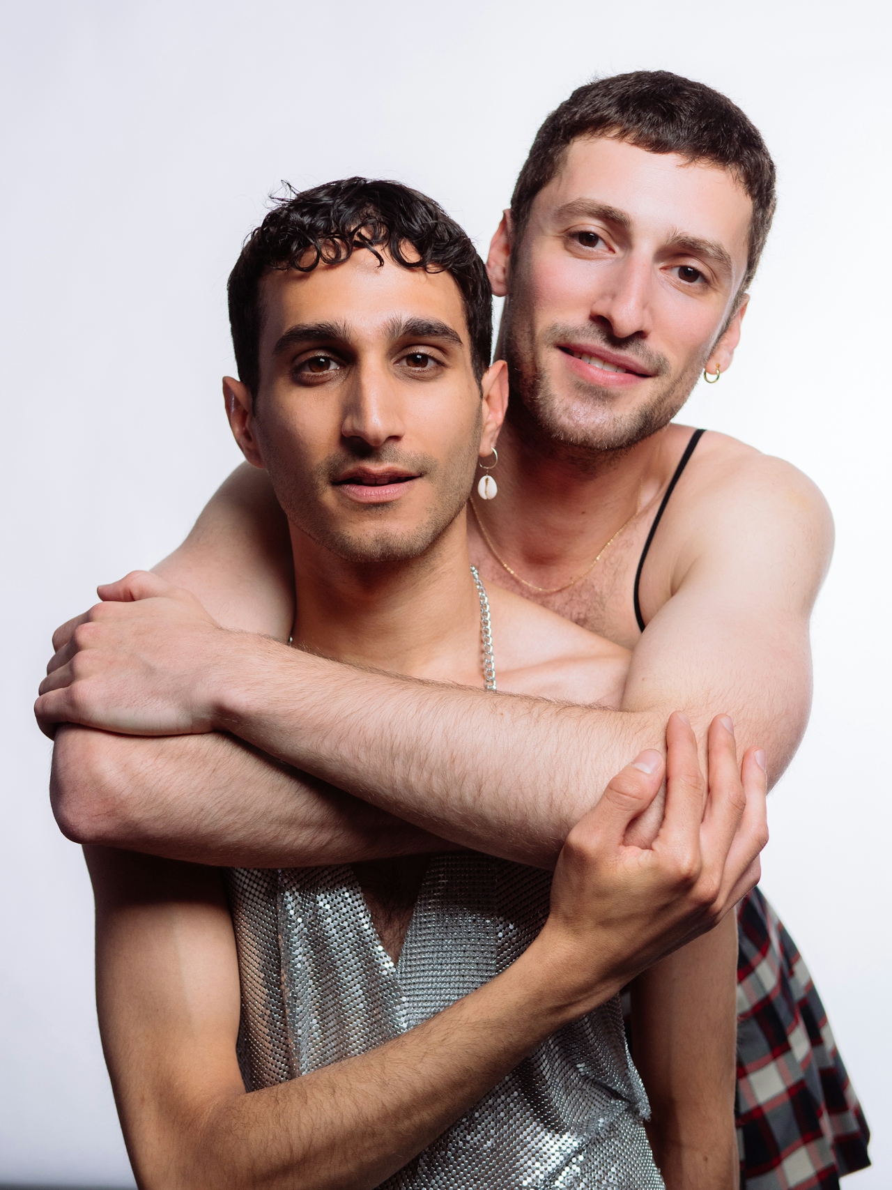 Two men embracing, one with short curly hair wearing a silver mesh top, the other with short hair and a plaid skirt, both smiling gently.