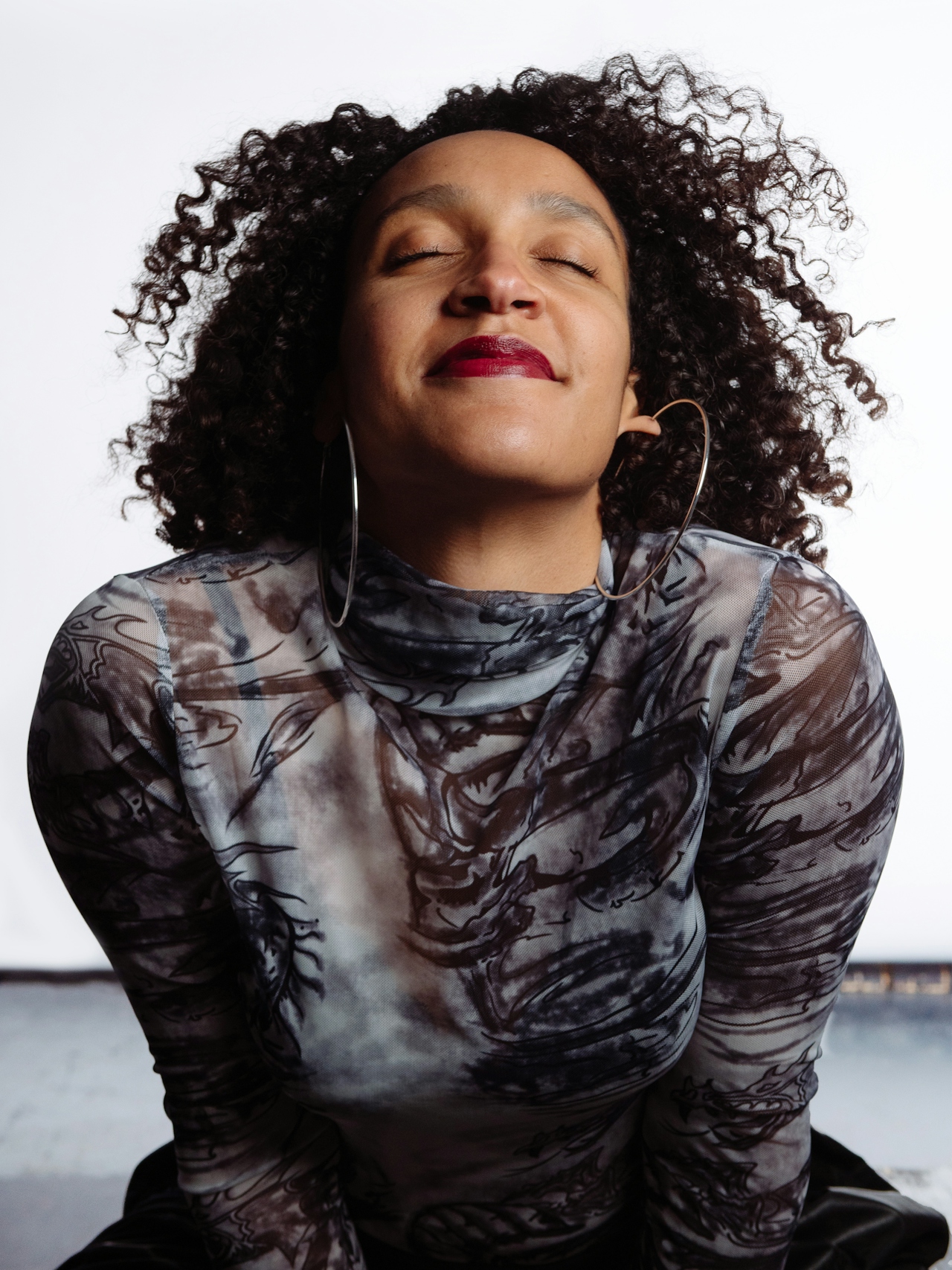 Person with curly hair, wearing large hoop earrings and a patterned long-sleeve top, smiling with eyes closed against a white background.