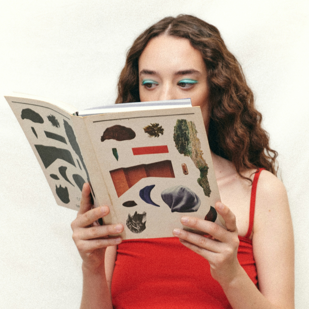 Young woman with curly hair and teal eyeshadow wearing a red tank top, reading a book with abstract art on the cover.