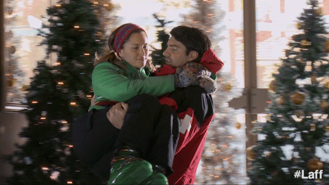 Man wearing a red jacket carrying a woman in a green jacket in front of decorated Christmas trees.