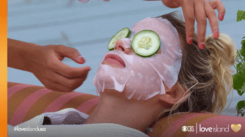Person lying down with a white facial mask and cucumber slices on their eyes while another person adjusts their hair.