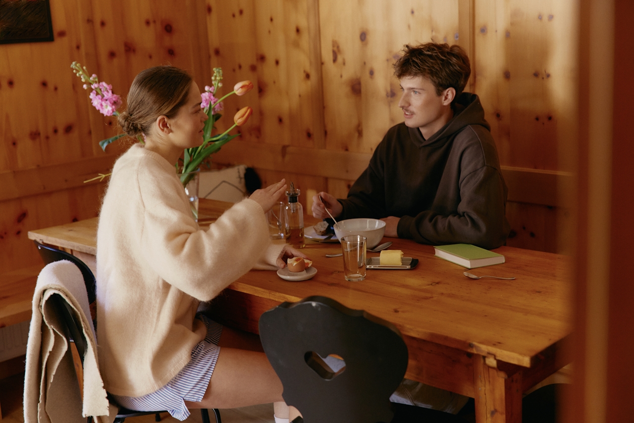 A young man and woman sitting at a wooden table indoors, having breakfast and talking, with flowers in a vase on the table.