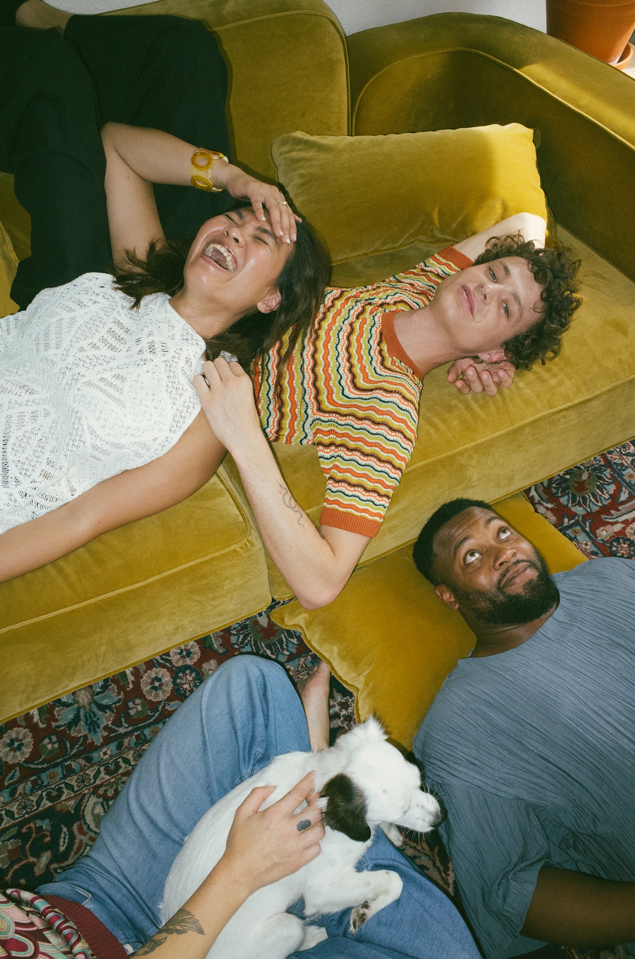 Four friends relaxing together on and around a green couch, one holding a small white dog, all appearing joyful and comfortable.
