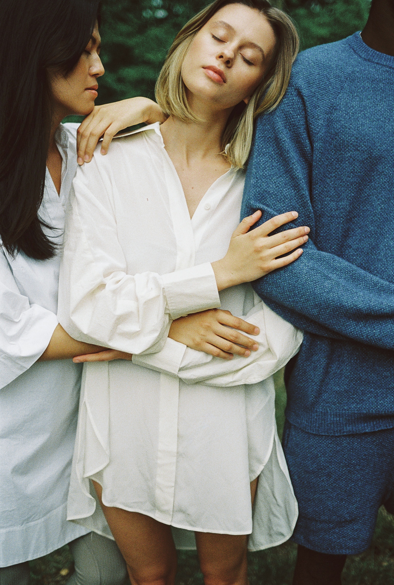 Three people closely embracing; a woman in the center with closed eyes wears a white shirt, leaning on a person in a blue sweater on the right, while another person on the left gently rests an arm on her shoulder.