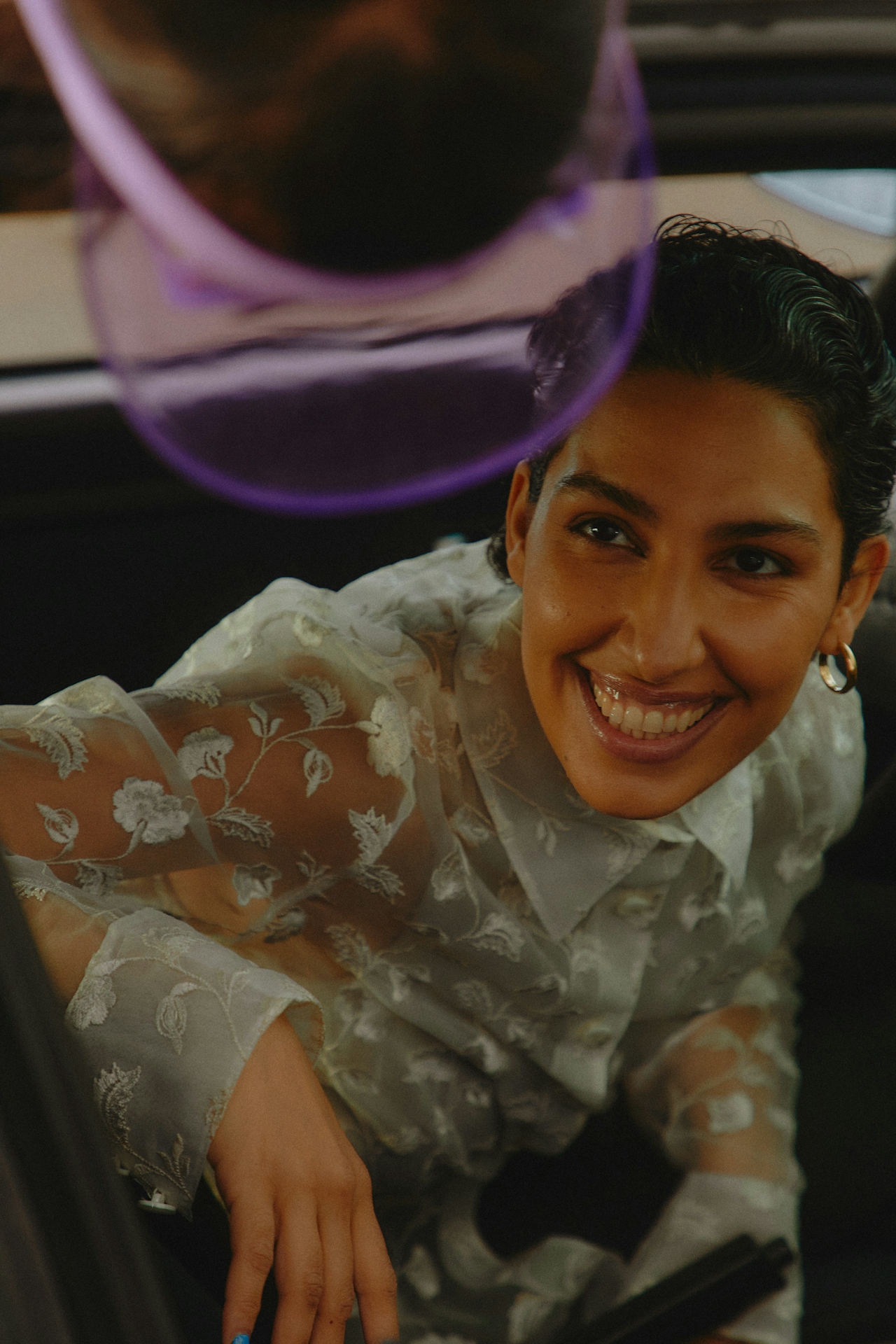Smiling woman with short dark hair and gold hoop earrings wearing a sheer floral embroidered blouse.