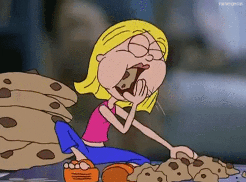 Animated girl with blonde hair sitting cross-legged and eagerly eating a chocolate chip cookie with a large stack of cookies beside her.