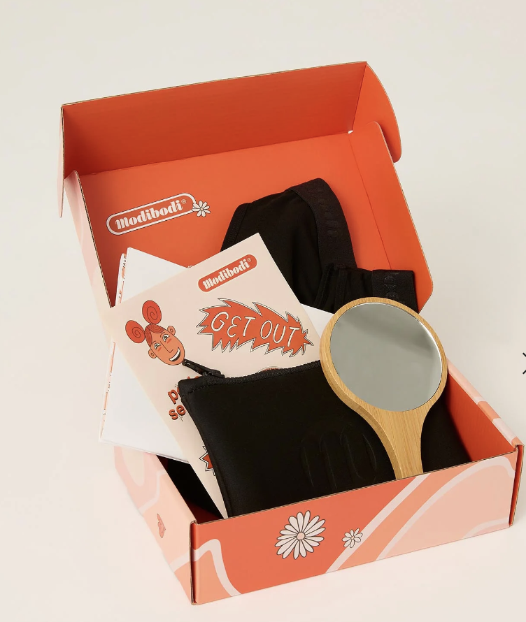 Open coral box containing a black pouch, a wooden handheld mirror, black fabric, and colorful cards with text.