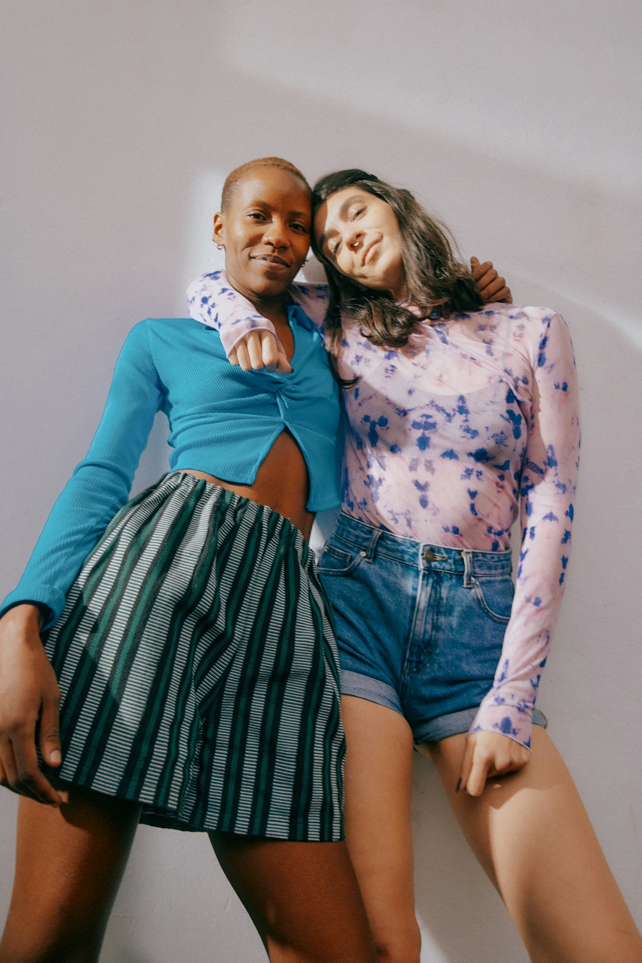Two women standing close together, one with short hair wearing a blue top and striped skirt, the other with long dark hair wearing a pink and blue patterned long-sleeve top and denim shorts.